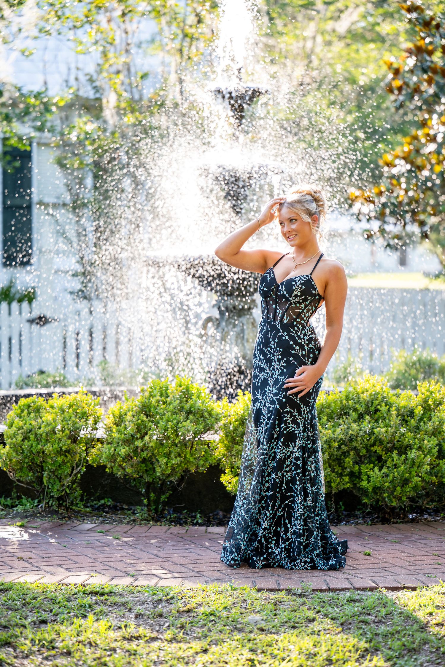 Woman in elegant navy sequin gown posing gracefully in front of a decorative outdoor fountain surrounded by lush greenery.