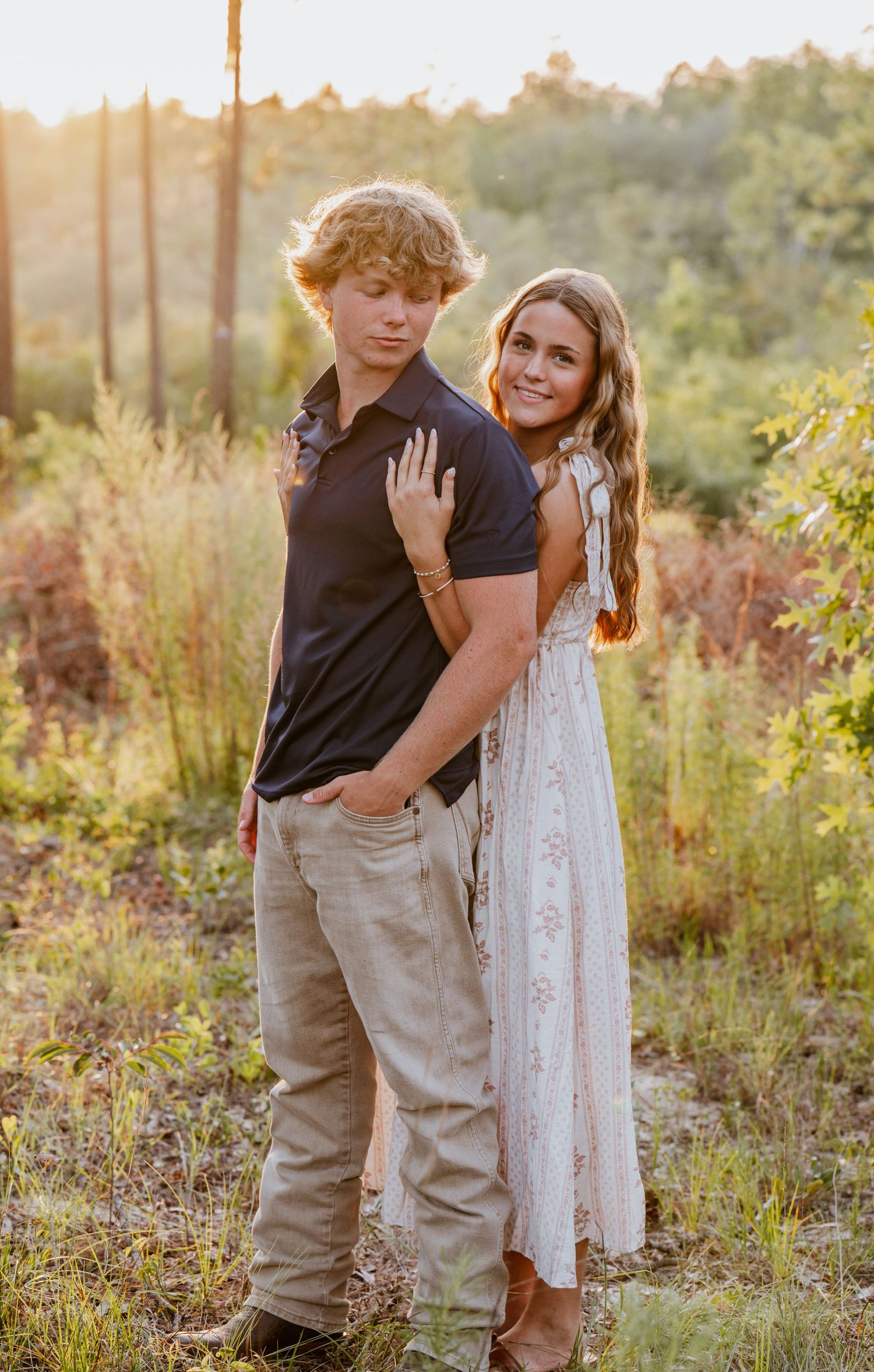 A young couple poses together in a golden-hour outdoor setting, surrounded by lush greenery.