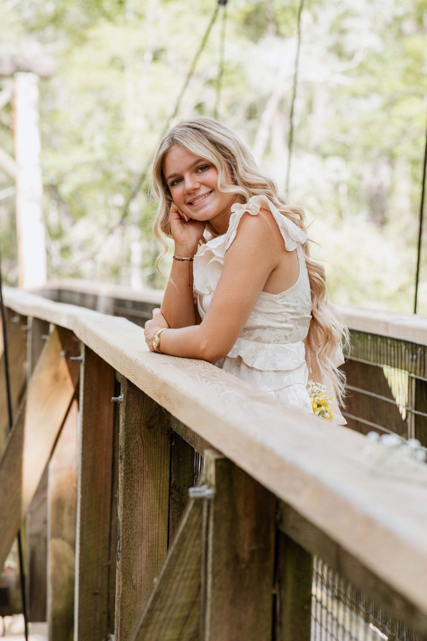 Smiling blonde woman in white ruffled dress leaning on wooden bridge railing surrounded by lush green trees.