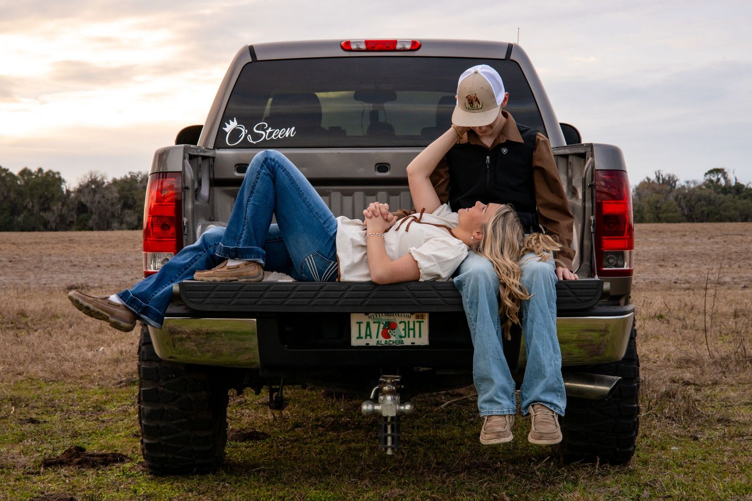 A couple relaxes together in the bed of a pickup truck at sunset in a rural field.