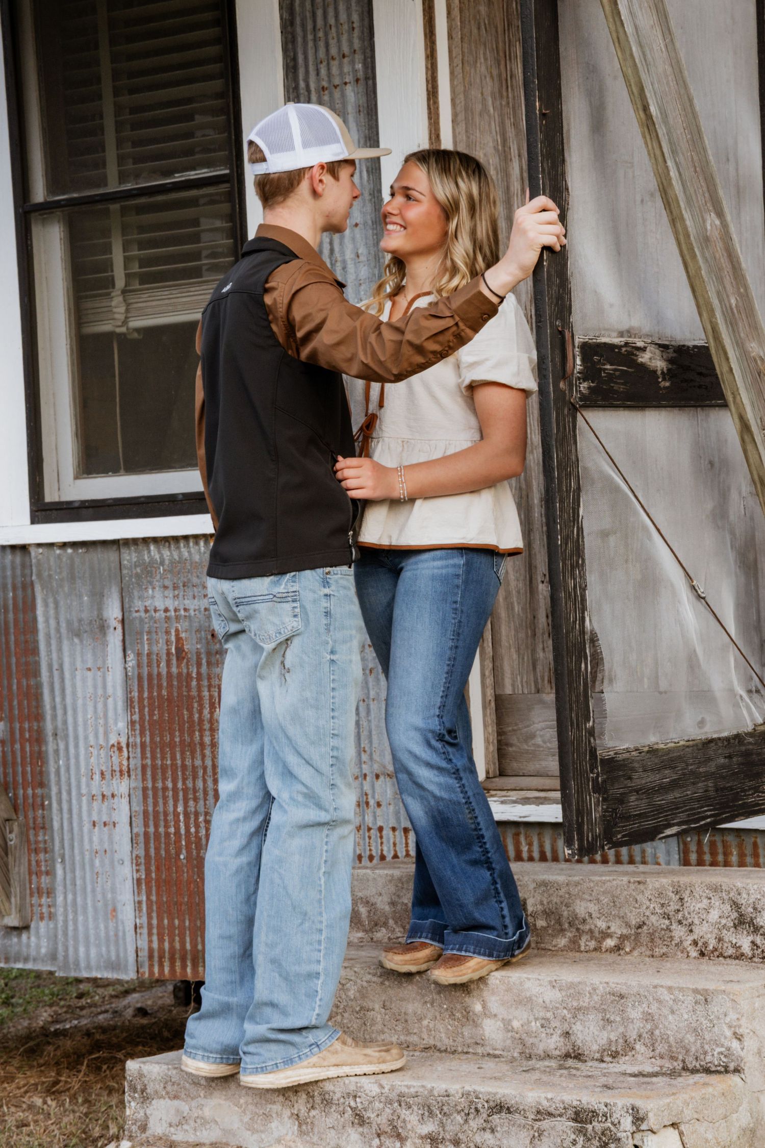 Couple in casual denim and white shirts embrace while standing on rustic wooden porch steps of old building.