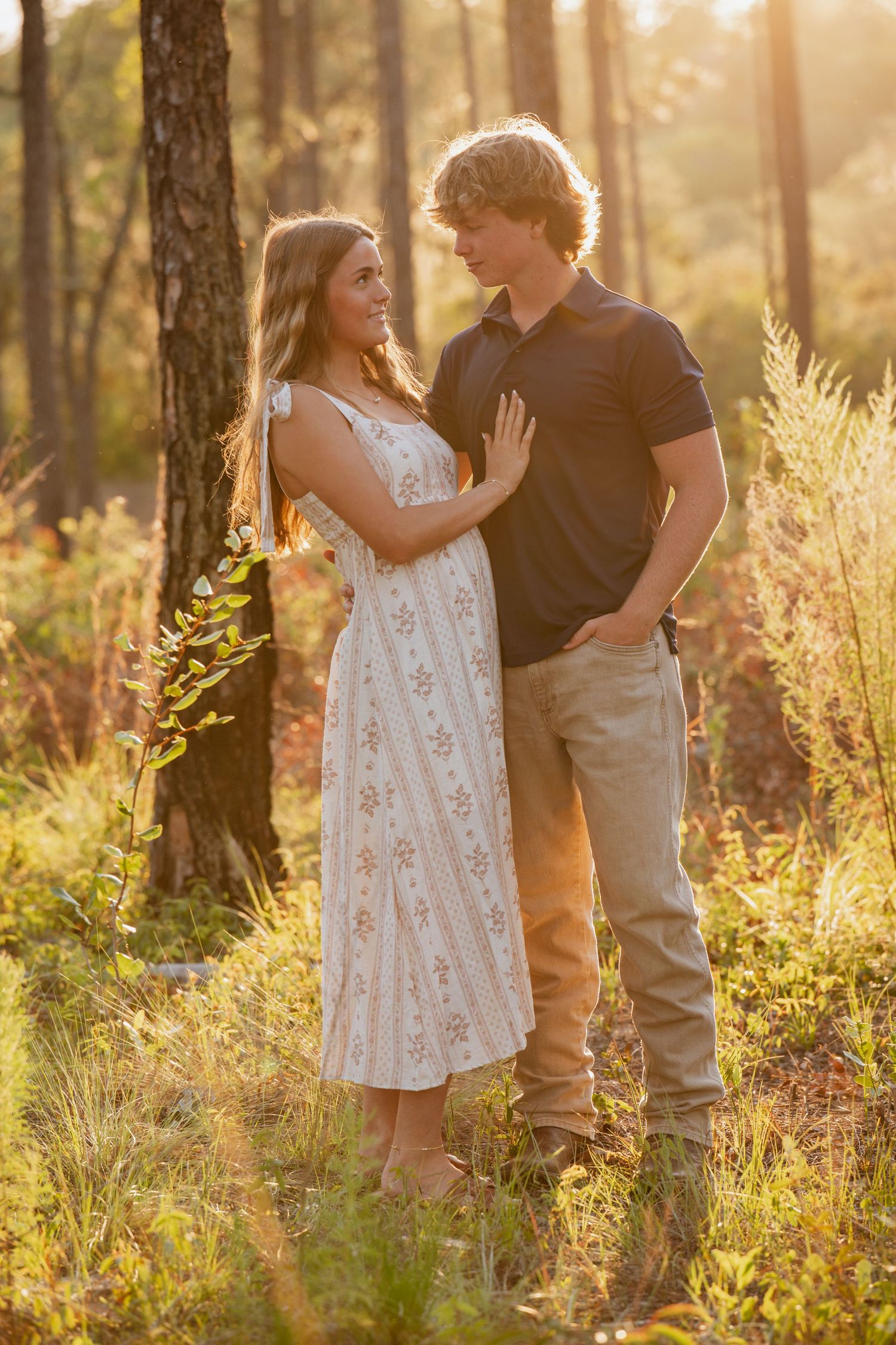 A young couple shares a romantic moment in a sunlit forest, she in a floral dress, he in a black polo shirt.