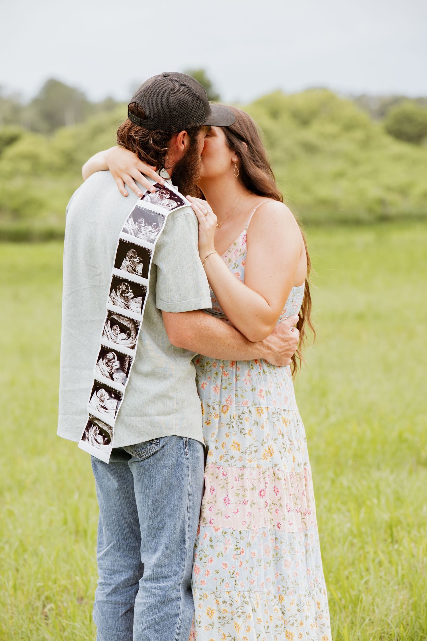 Couple kissing in green field holding ultrasound photos, announcing pregnancy in floral dress and cap outdoors.