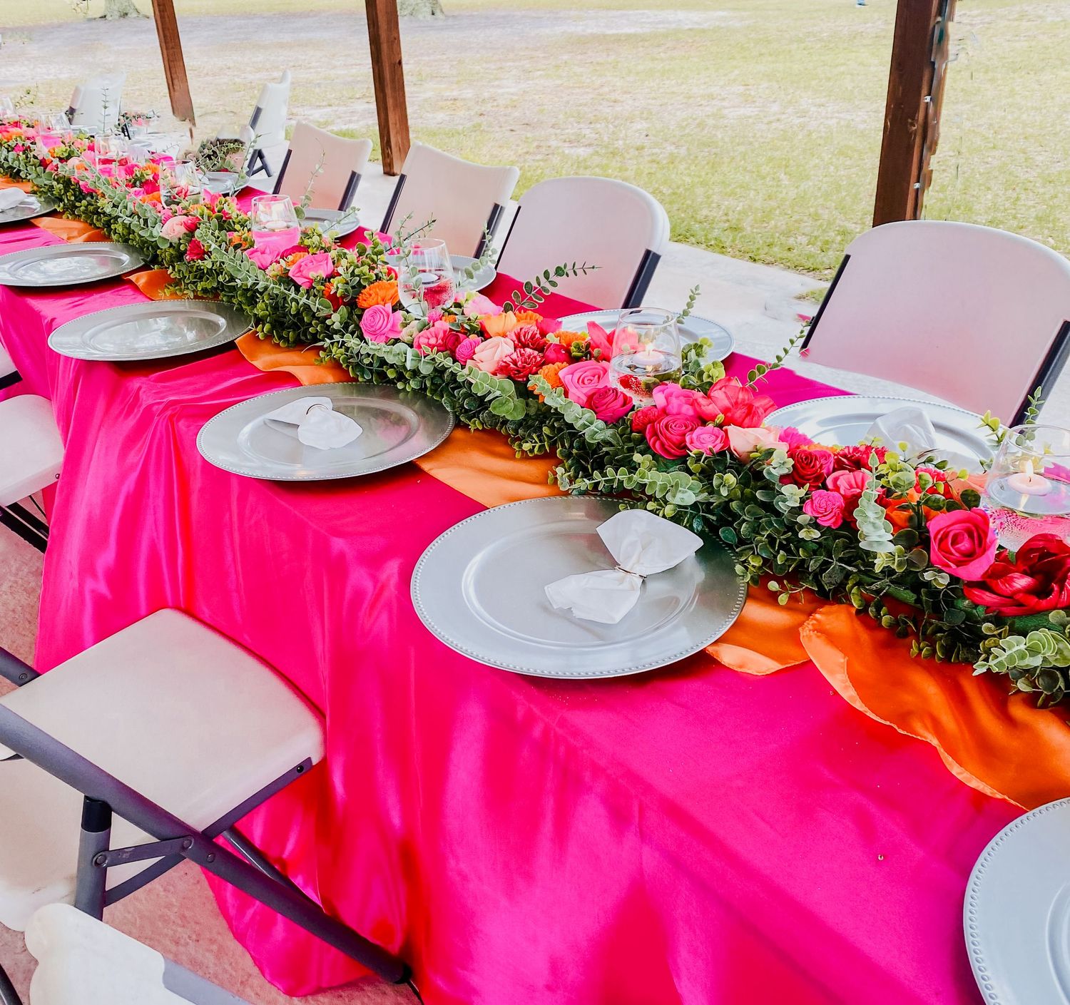 Elegant long banquet table with hot pink tablecloth, floral garland centerpiece, and gray charger plates at an outdoor event.