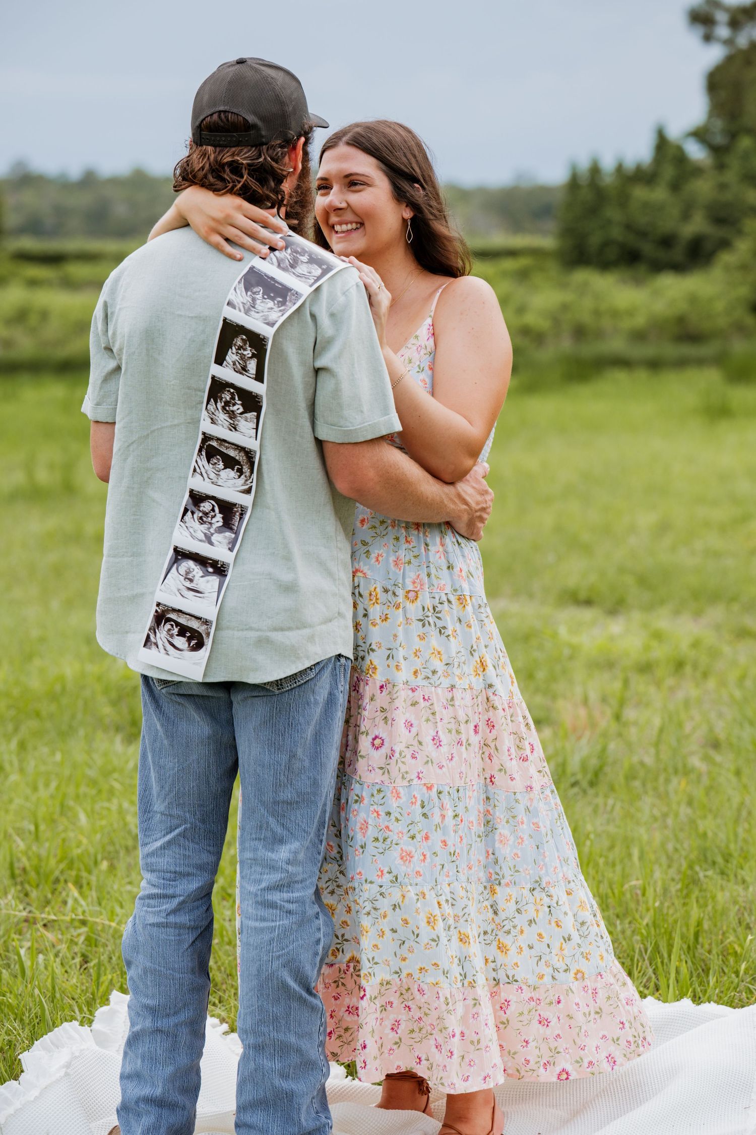 Couple embracing outdoors holding ultrasound photos, woman in floral dress smiling, pregnancy announcement in green field.
