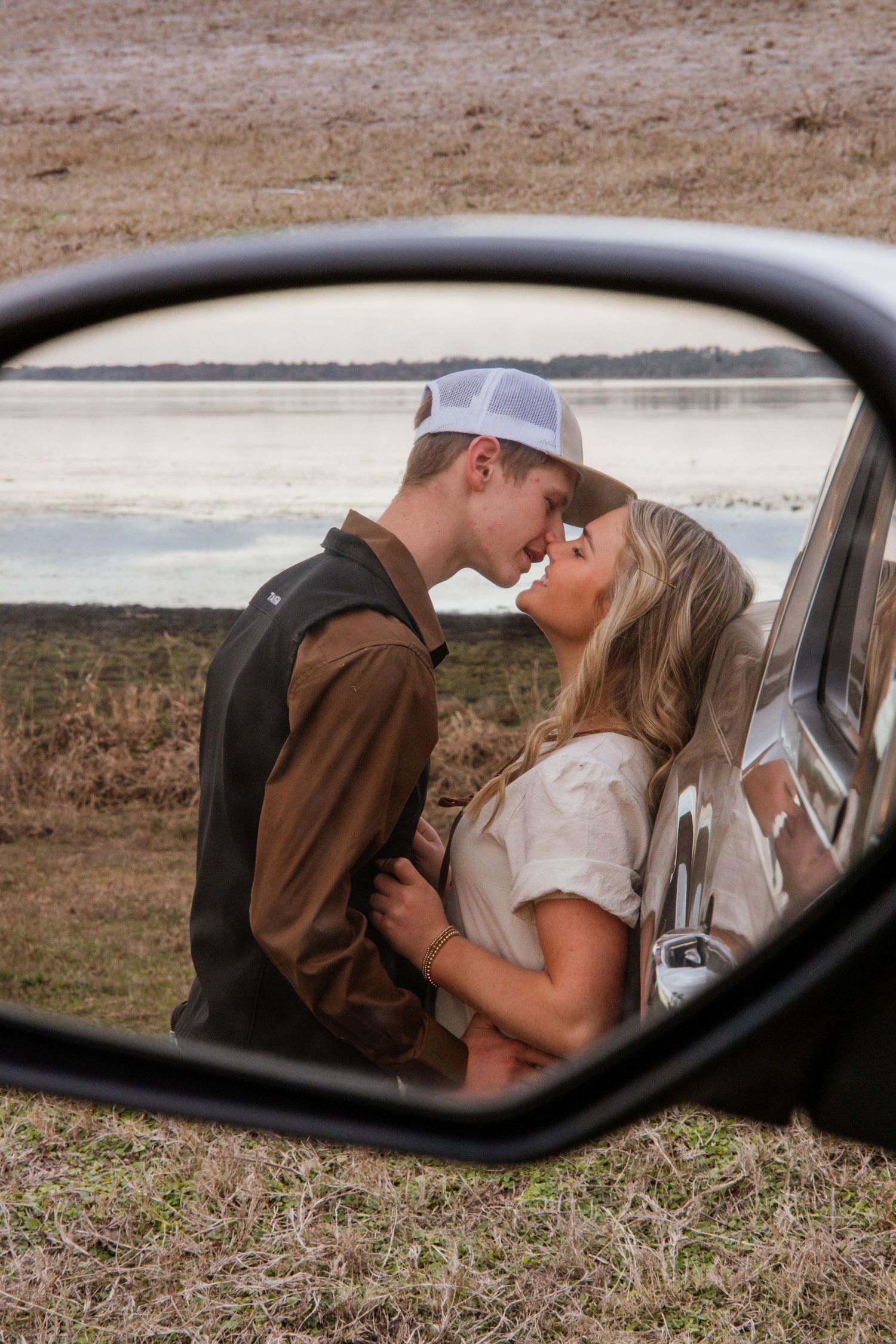 Romantic couple embraces near vintage car, viewed through side mirror reflection against rural landscape at sunset.
