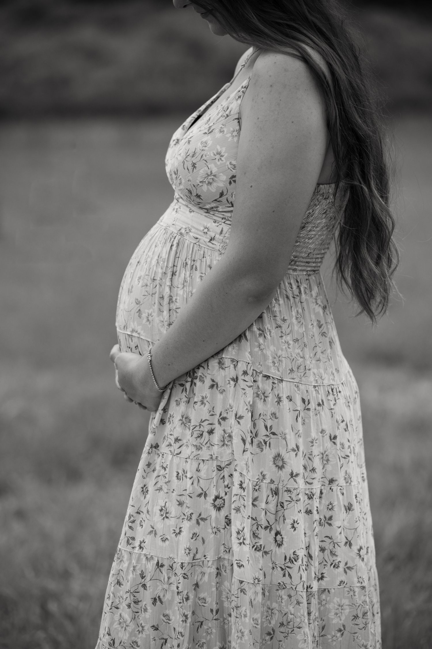Black and white photo of a pregnant woman in a floral dress cradling her baby bump outdoors.
