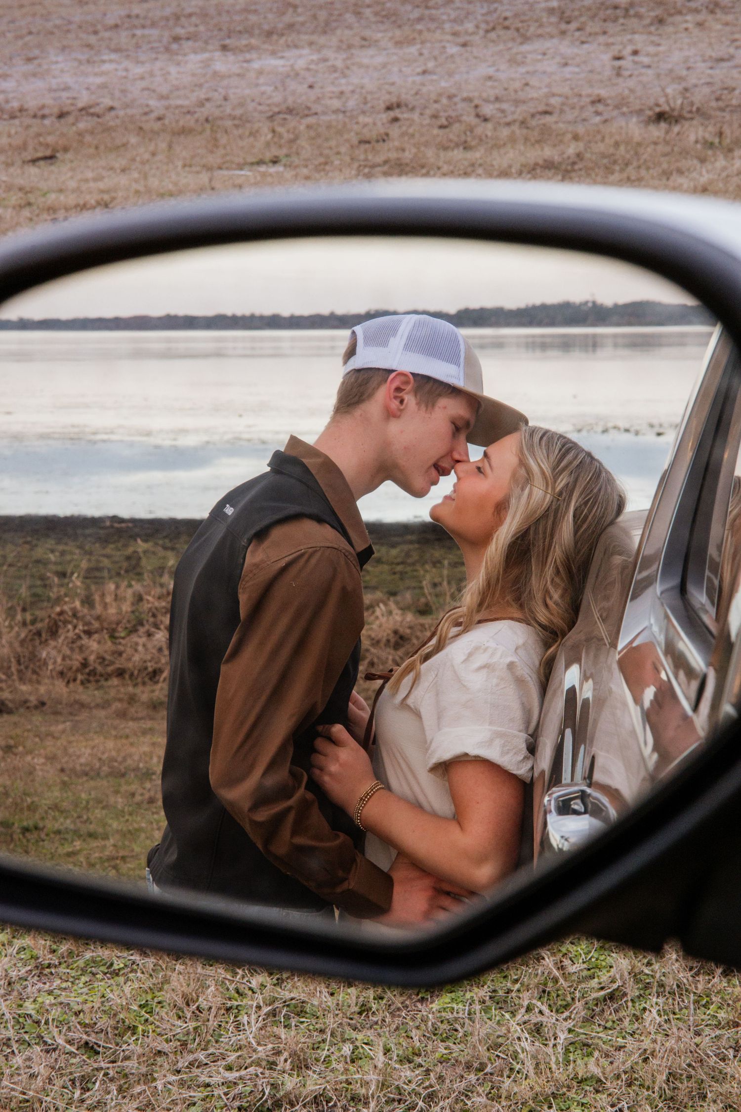 Couple sharing a romantic moment reflected in a truck side mirror near a serene lake backdrop.