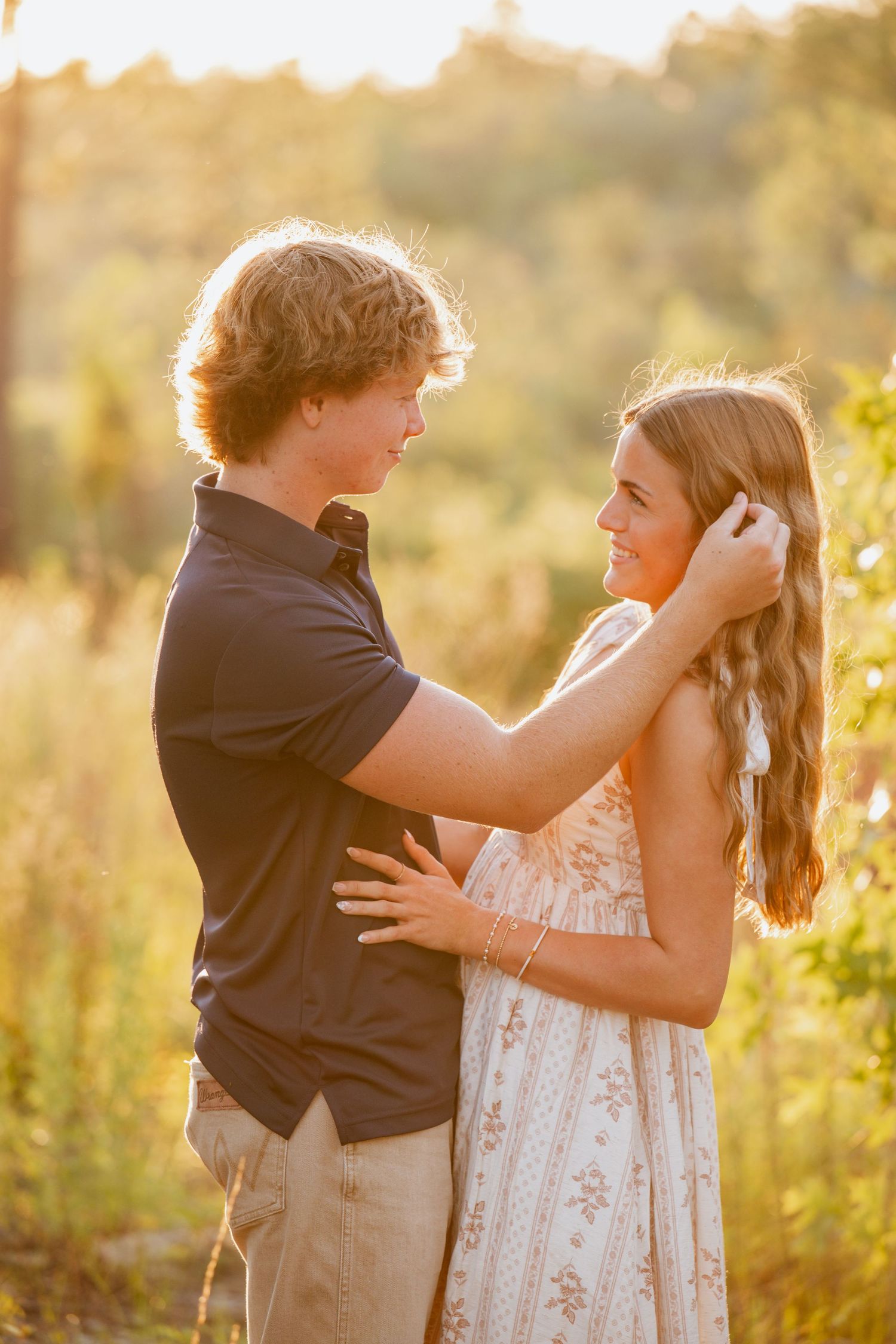 A young couple shares a tender moment outdoors at golden hour, as he gently brushes her long hair back.