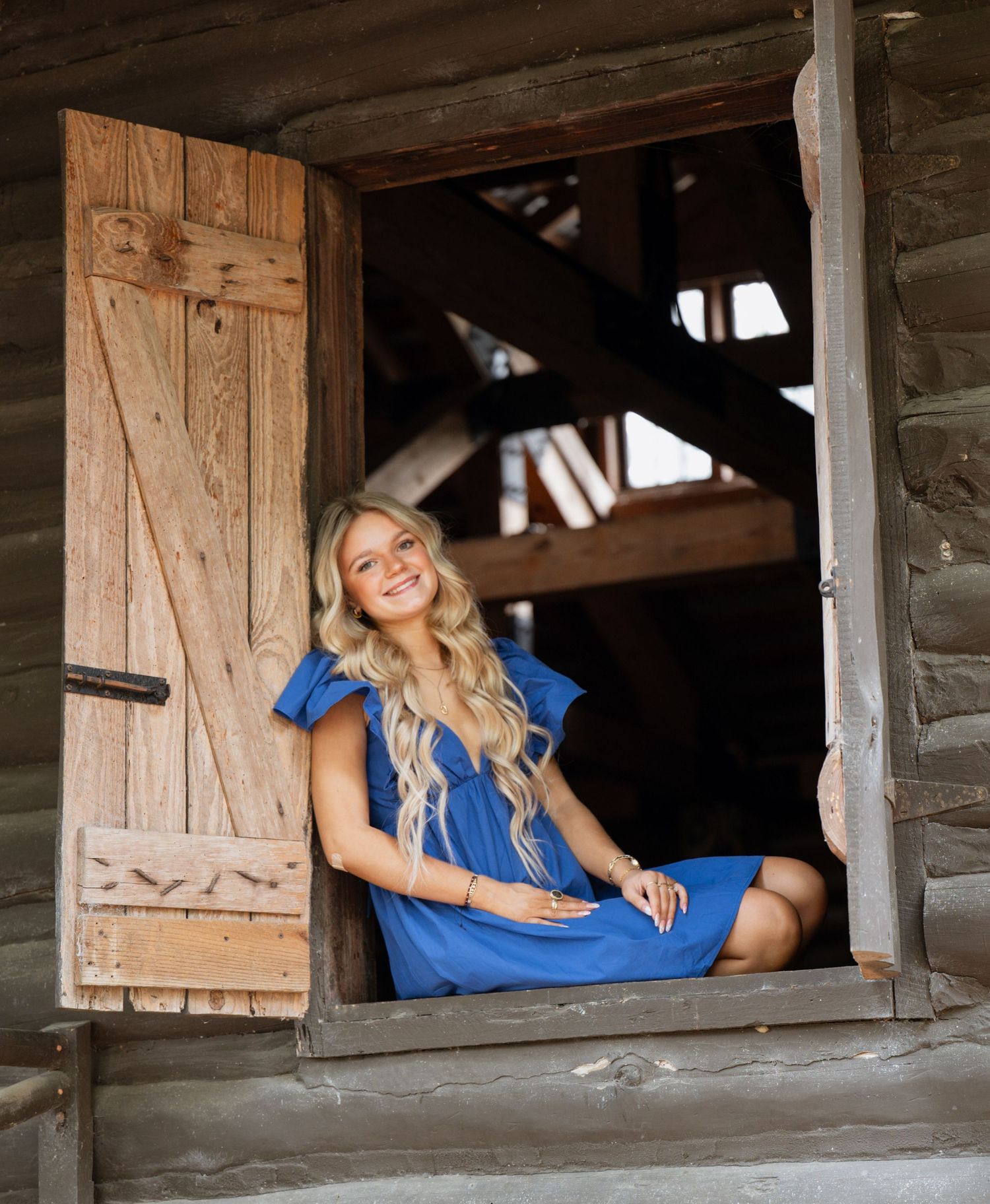 Young blonde woman in blue dress sitting in rustic wooden barn window, smiling brightly.