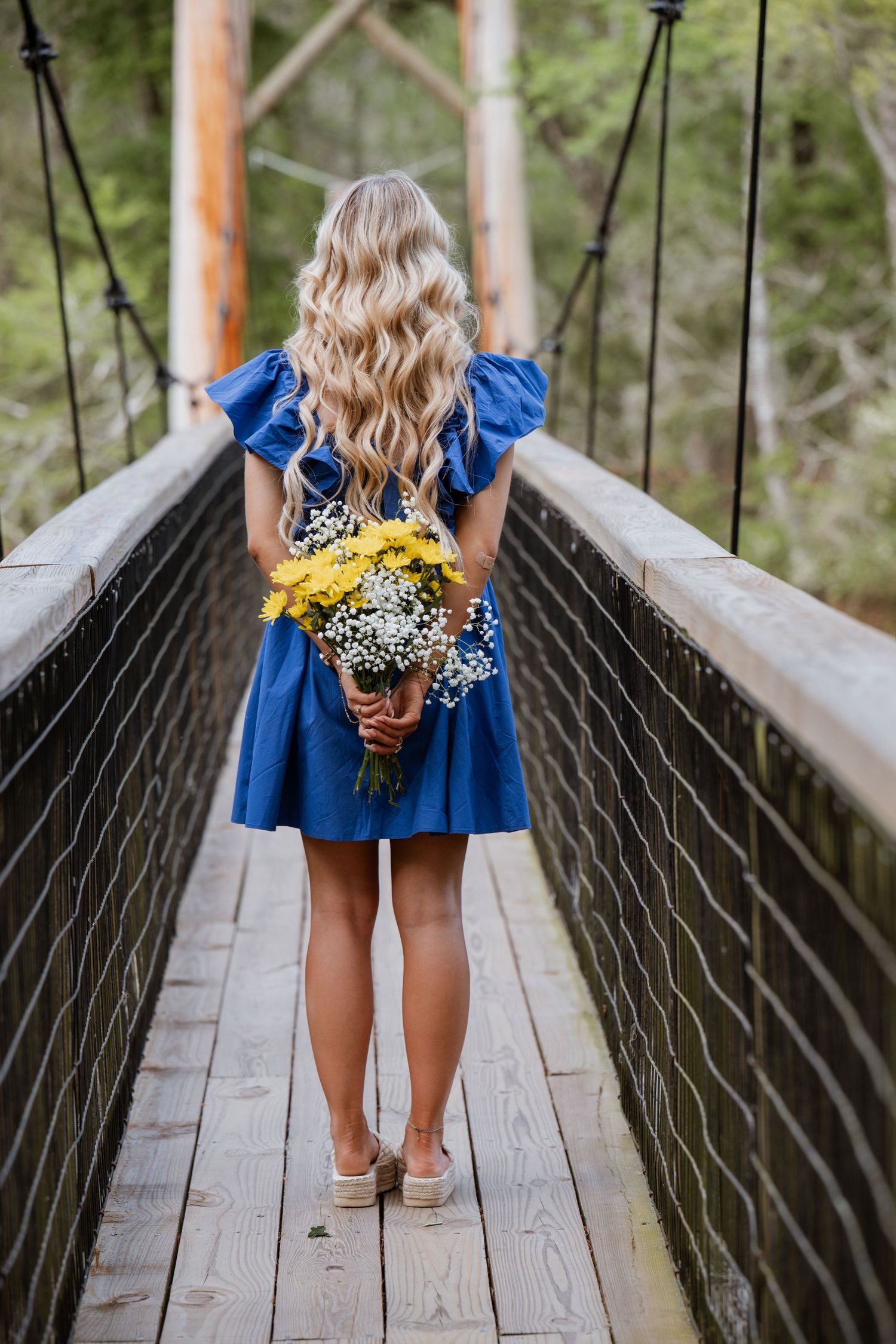 Blonde woman in blue dress holding yellow wildflowers stands on wooden suspension bridge surrounded by lush forest.