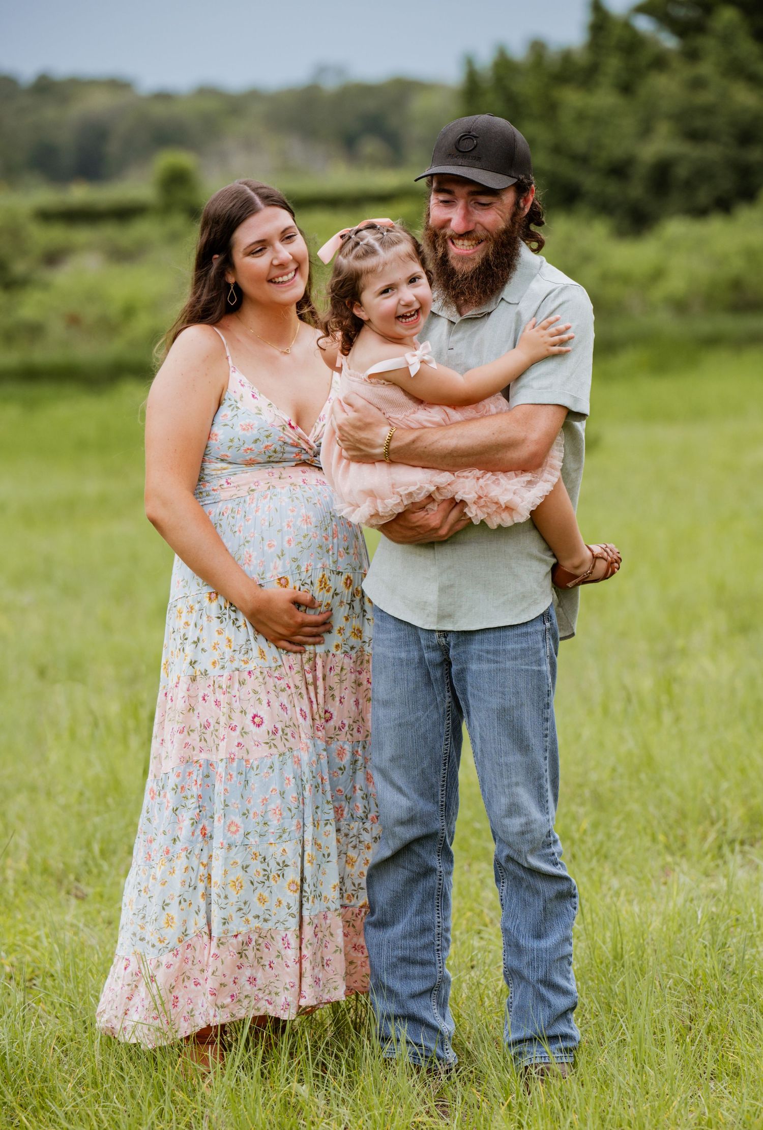 Pregnant woman in floral dress poses with bearded man in cap holding smiling toddler girl in pink tutu outdoors.