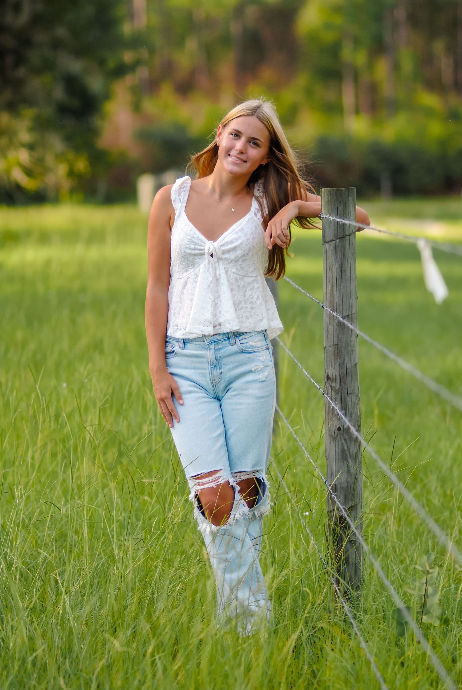 A person in distressed jeans and white top poses casually against a wooden fence post in a sunny green field.