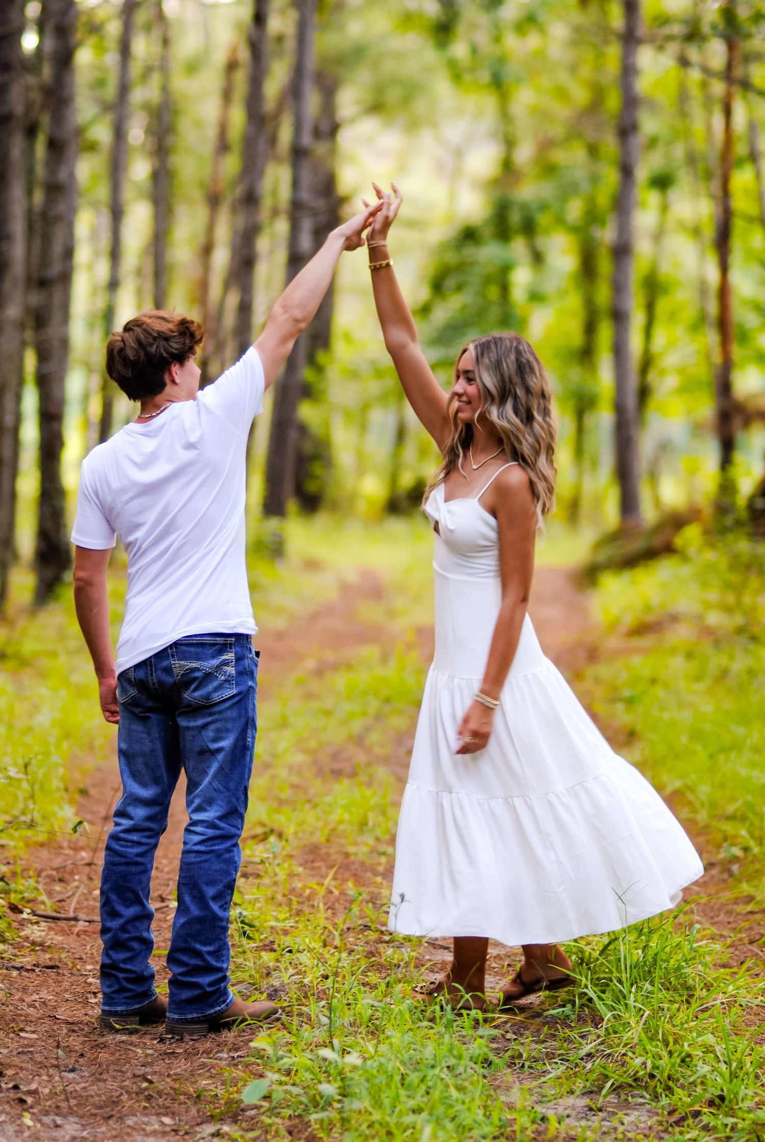 Two people raise their arms joyfully while walking down a tree-lined forest path.