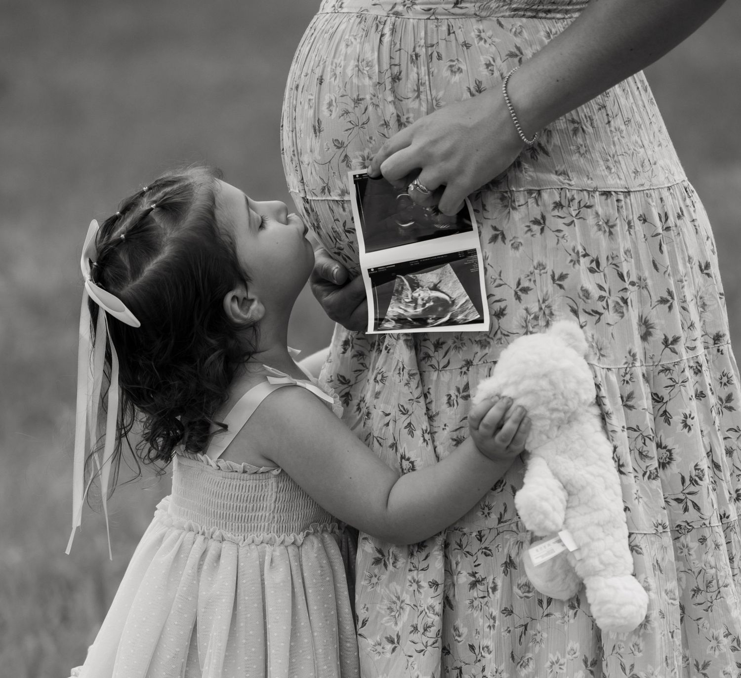 Black and white photo of a little girl kissing her pregnant mother's belly while holding a teddy bear and ultrasound photo.
