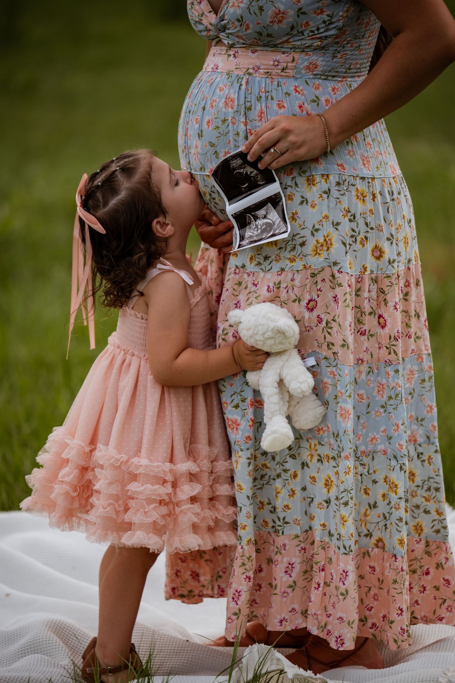 A young girl in a pink dress kisses her pregnant mother's belly while holding a stuffed bunny and ultrasound photos outdoors.