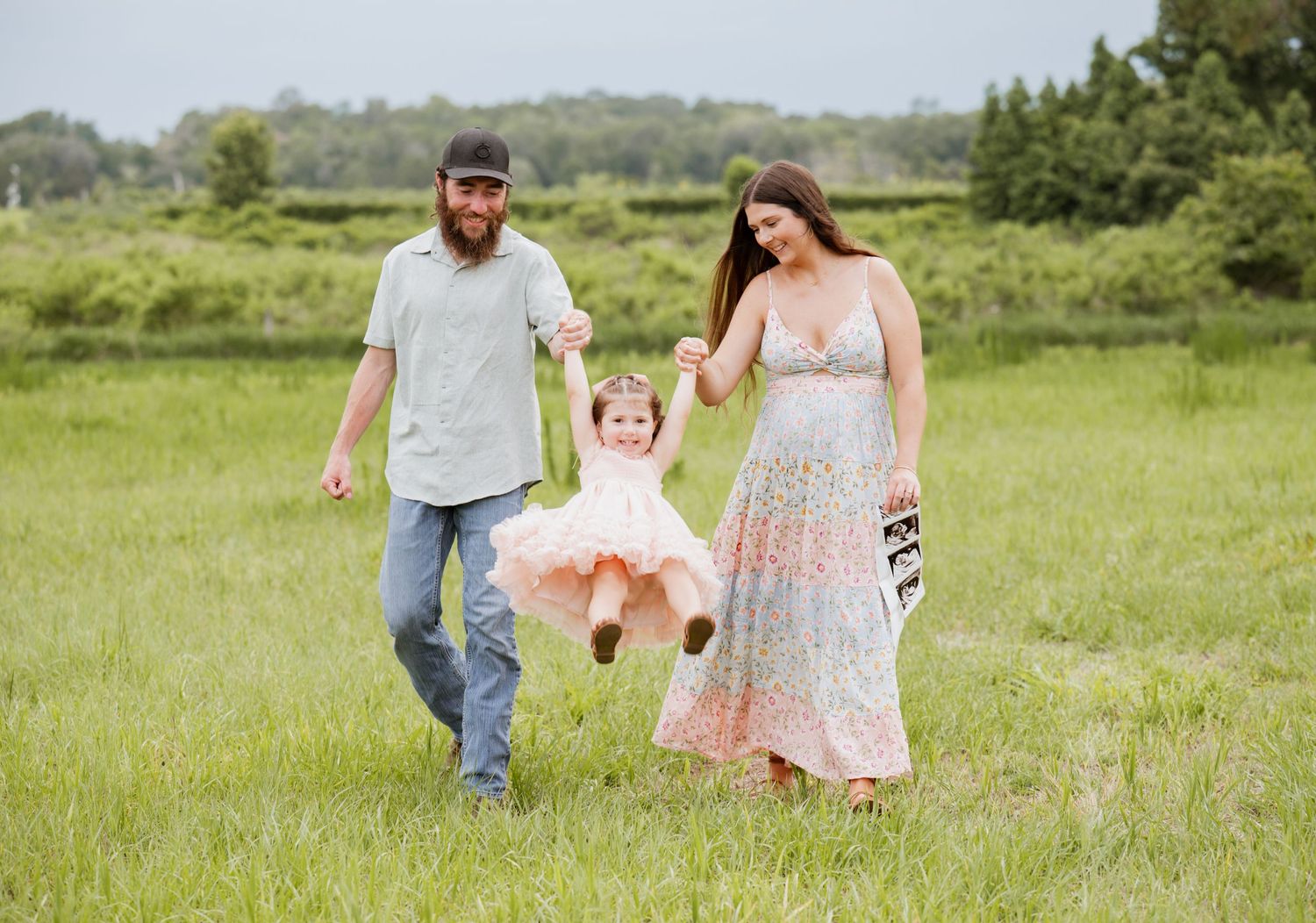 A happy family swings their toddler girl in a green meadow, mom in floral dress, dad in casual wear.