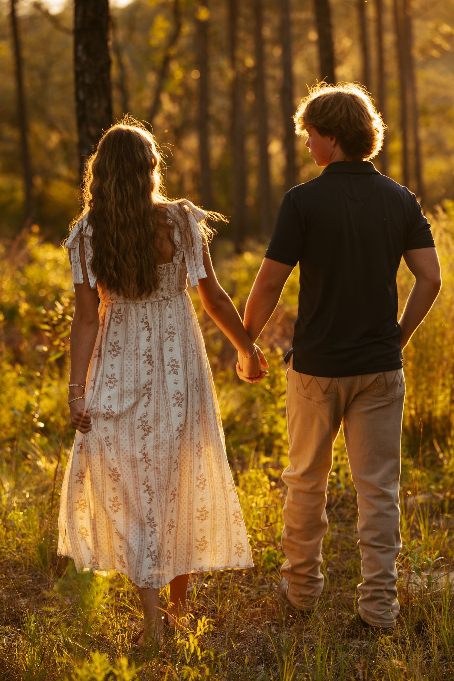 A couple holds hands while walking through a sunlit forest at golden hour, the woman in a floral dress and man in black polo.