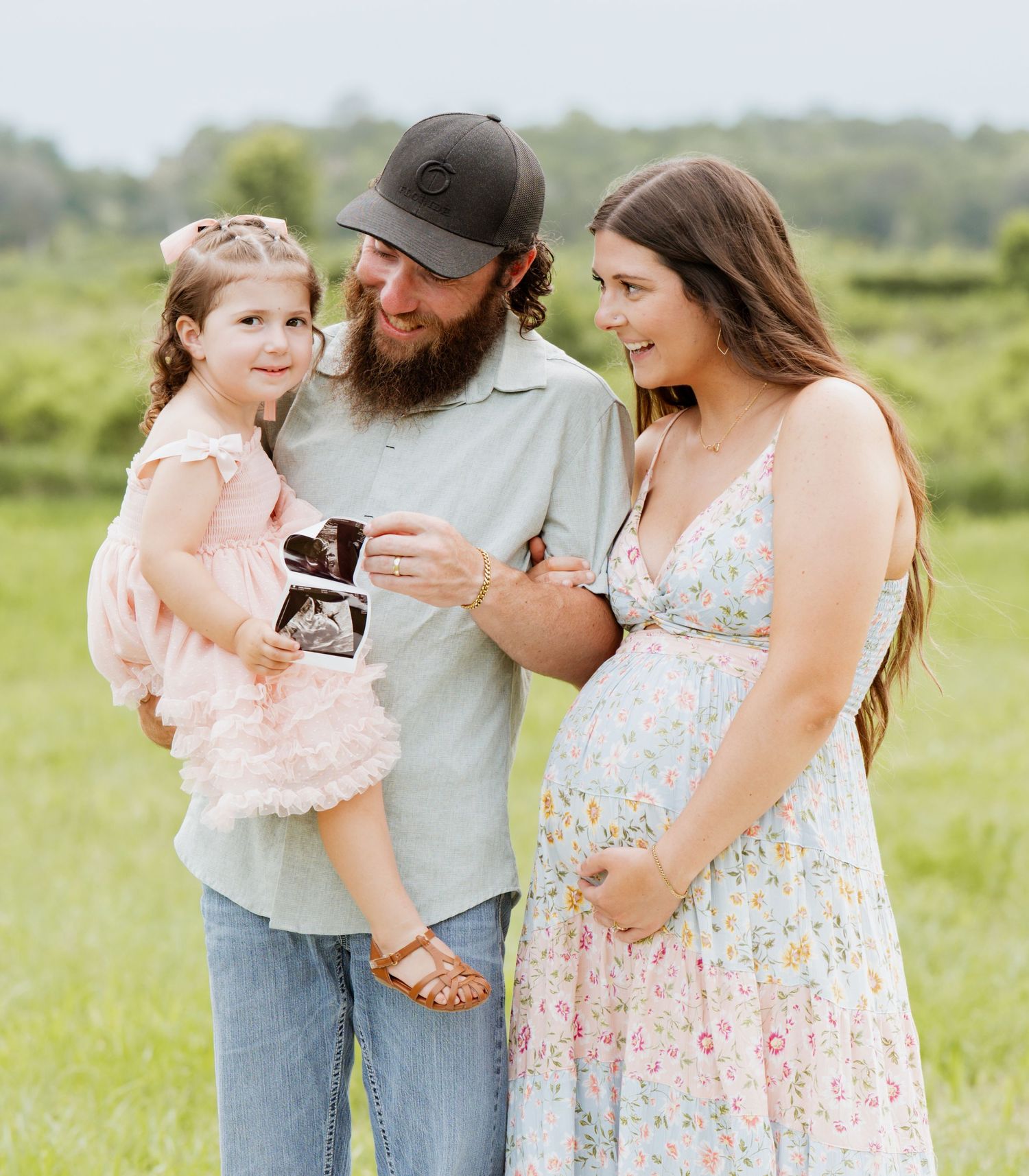 Pregnant woman in floral dress with bearded man in cap holding toddler girl displaying ultrasound photo outdoors.