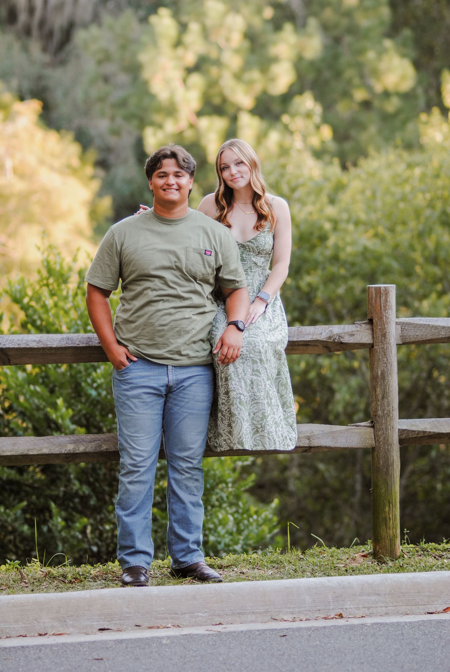 A couple poses casually along a wooden fence in a park setting.
