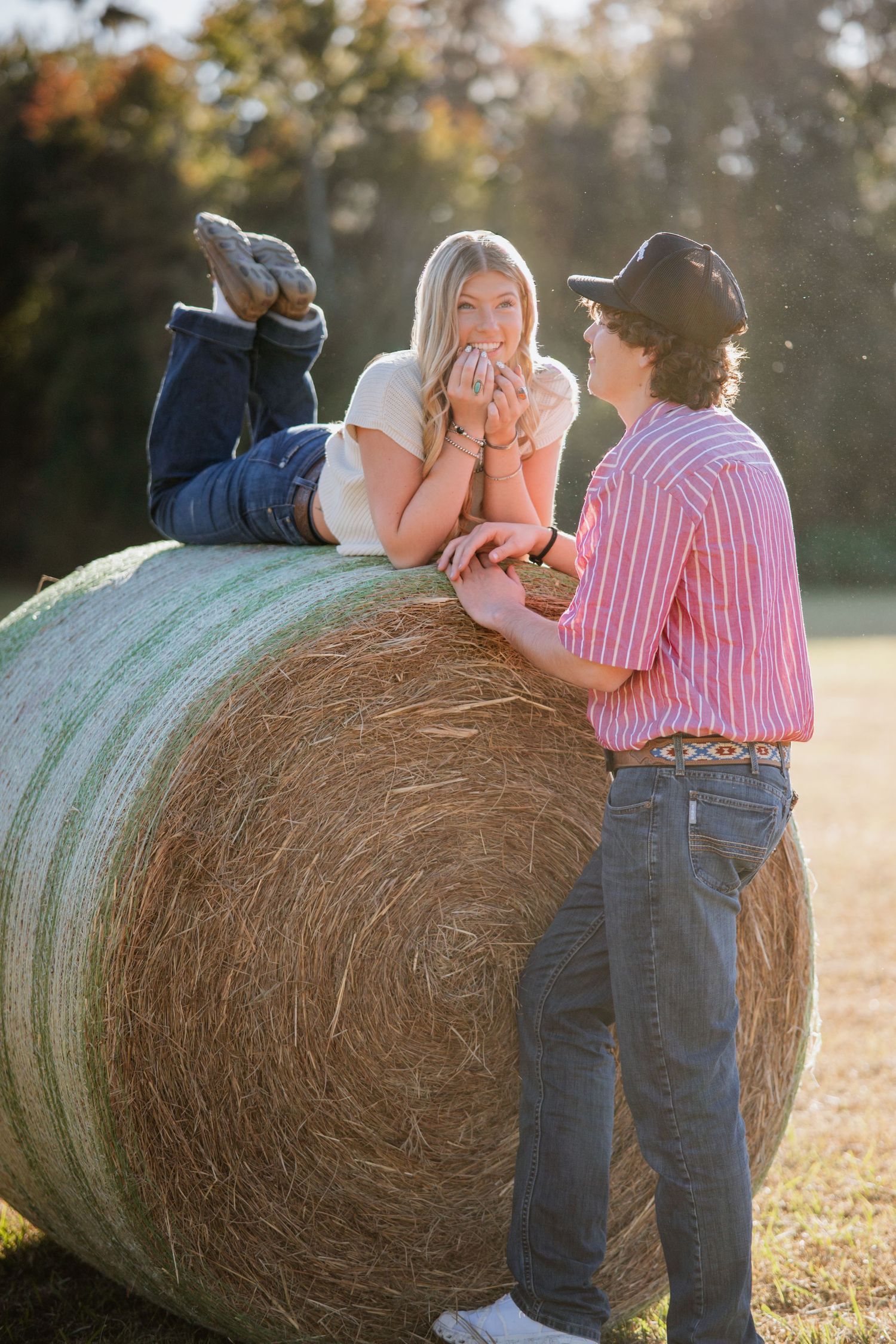 Two people sit playfully on a large round hay bale in a sunny field during sunset.