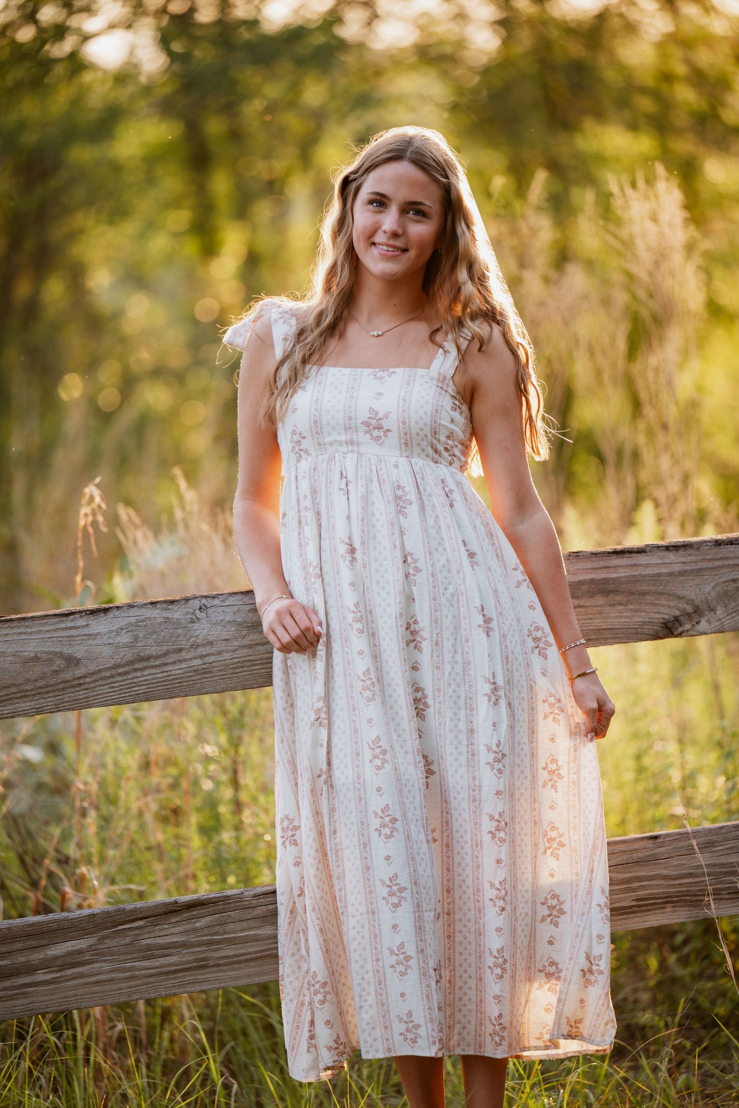 Young woman in floral midi dress poses by rustic wooden fence in golden hour sunlight outdoors.