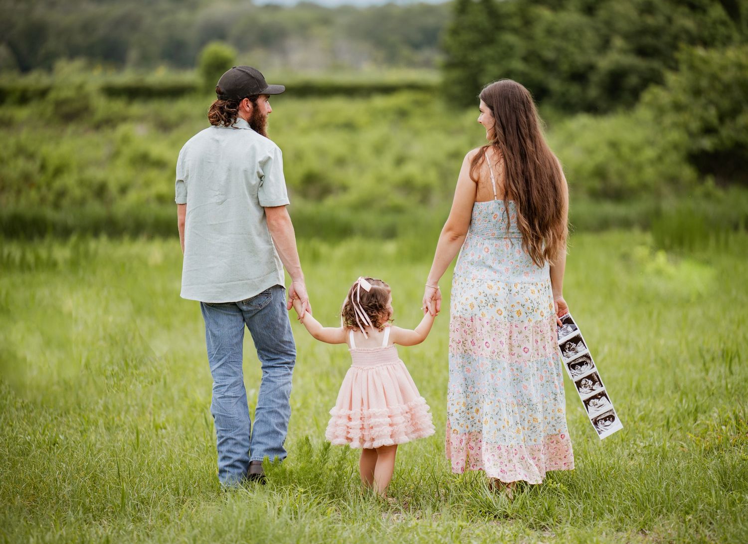 Family of three holding hands in green field, woman holding ultrasound photos, pregnancy announcement outdoor photo.