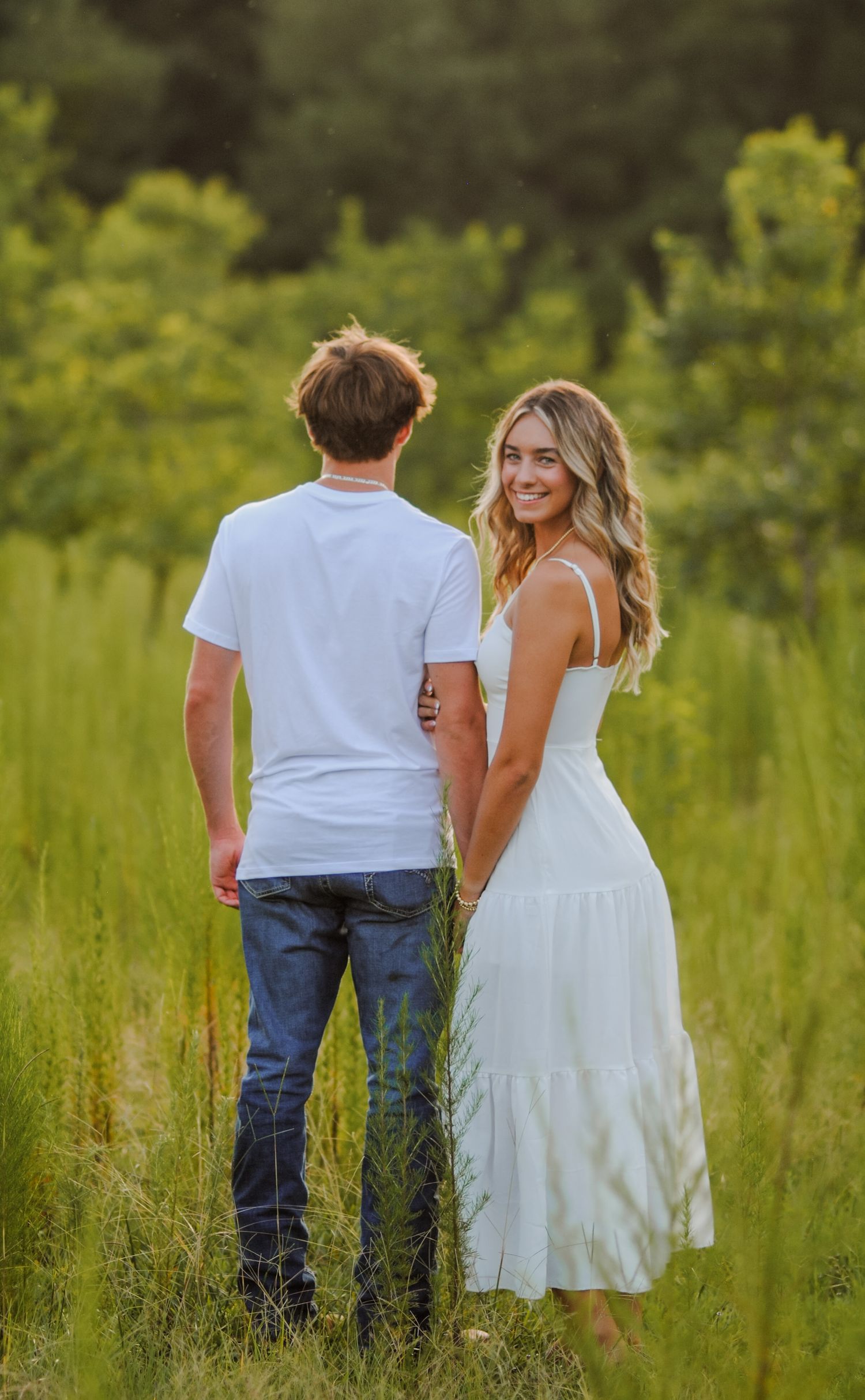 A couple walks through tall grass in a meadow with soft evening light filtering through.