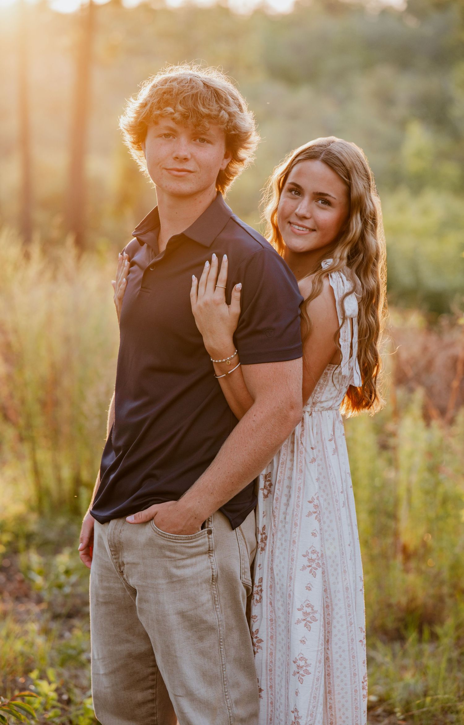 A young couple poses together at golden hour in a field, the girl in a floral dress resting her hand on the boy's back.