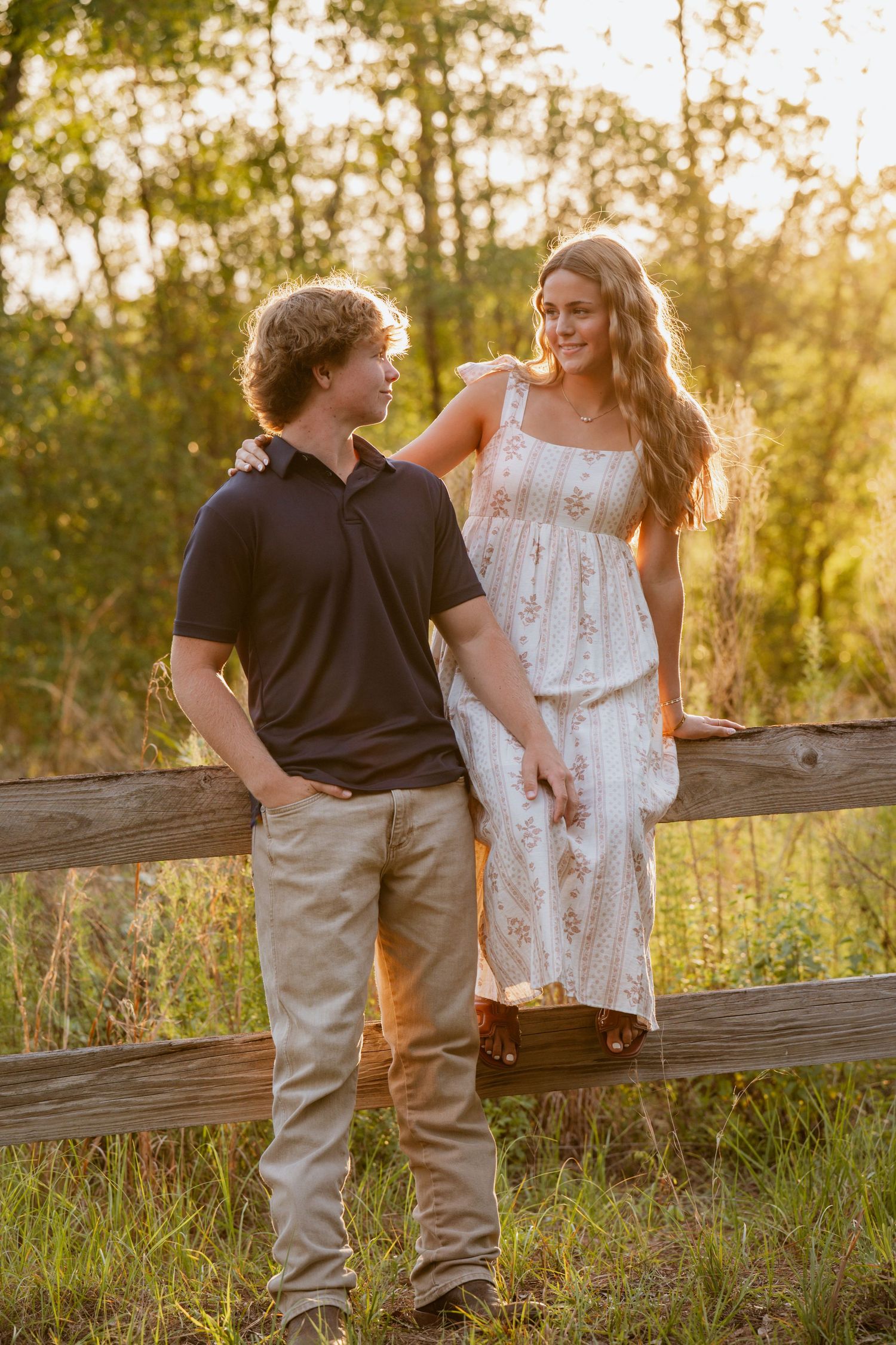 A young couple shares a romantic moment by a rustic wooden fence in a golden sunset field.