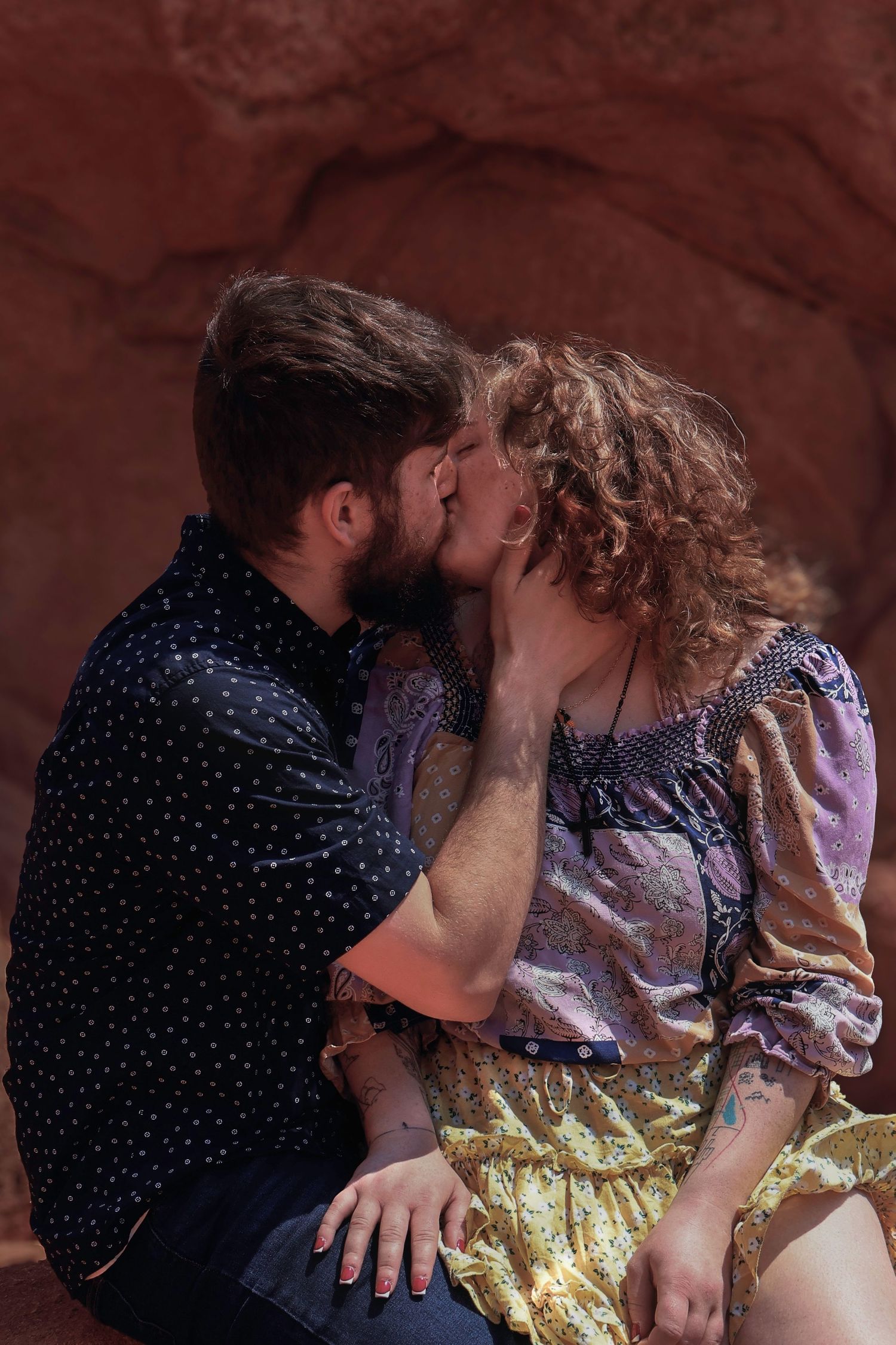 A couple sharing a romantic kiss while sitting against a red rock formation outdoors.