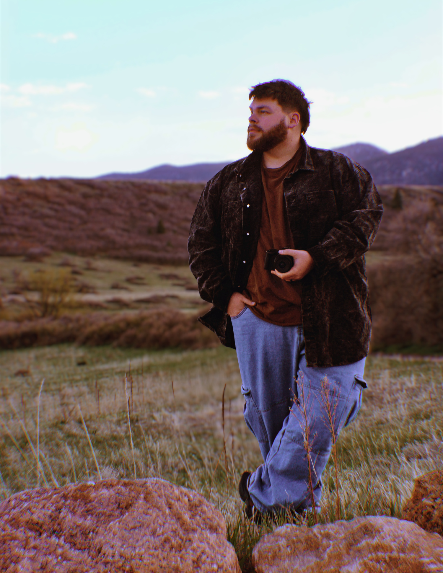Man in dark jacket and jeans standing on rocky hillside with mountains and open field in background at Forest Lakes in Monument, CO.