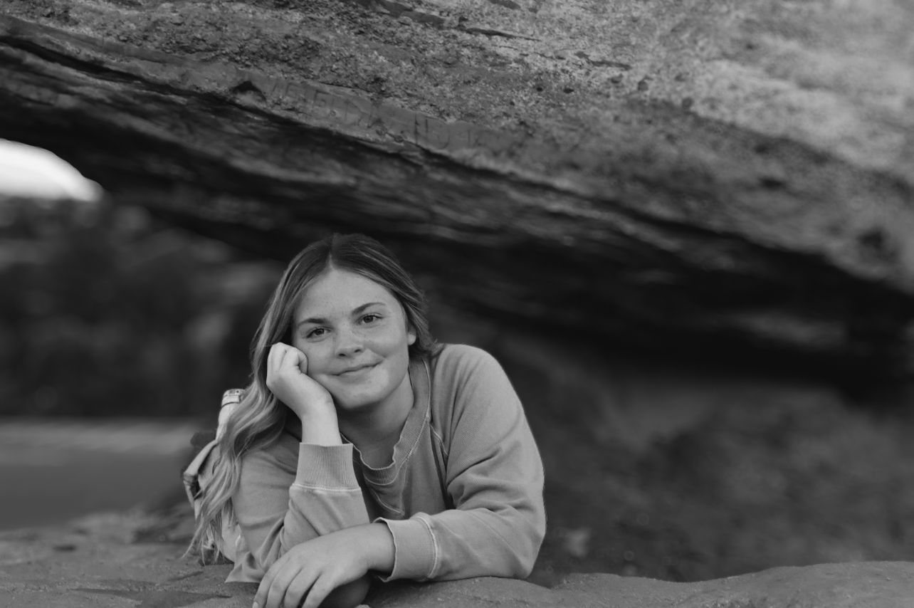 Girl smiles softly while leaning on a rock under natural stone arch. Outdoor black and white senior photo.