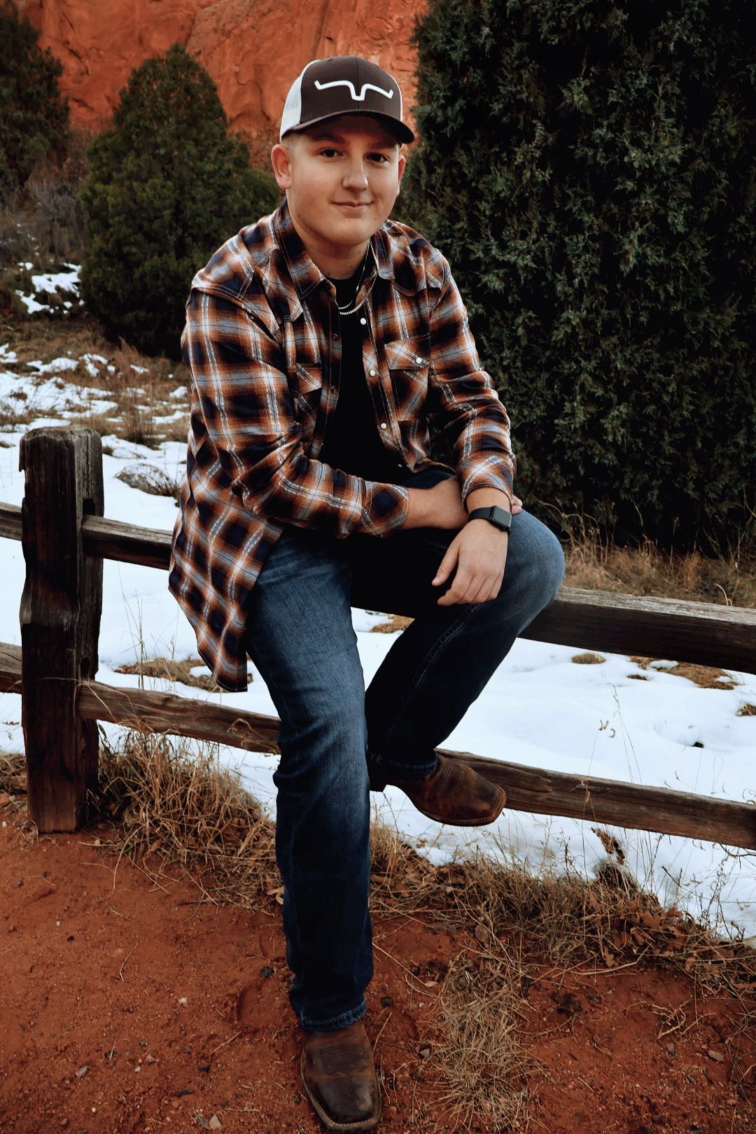 Teen boy in flannel shirt poses seated on a wooden fence with snowy ground. Rustic senior photo at Garden of the Gods.