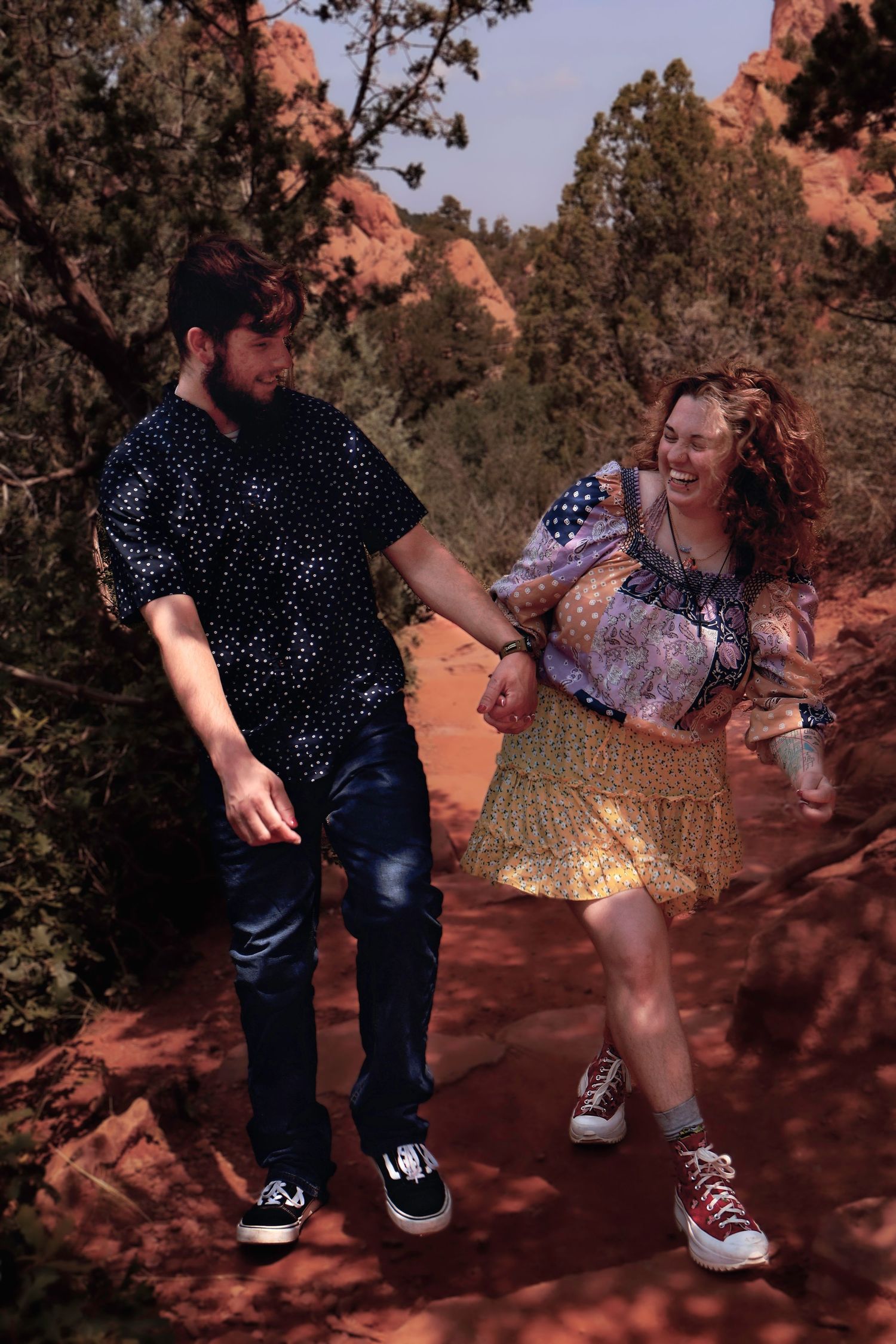Couple holding hands and laughing while hiking on a red dirt trail surrounded by trees and red rock formations.