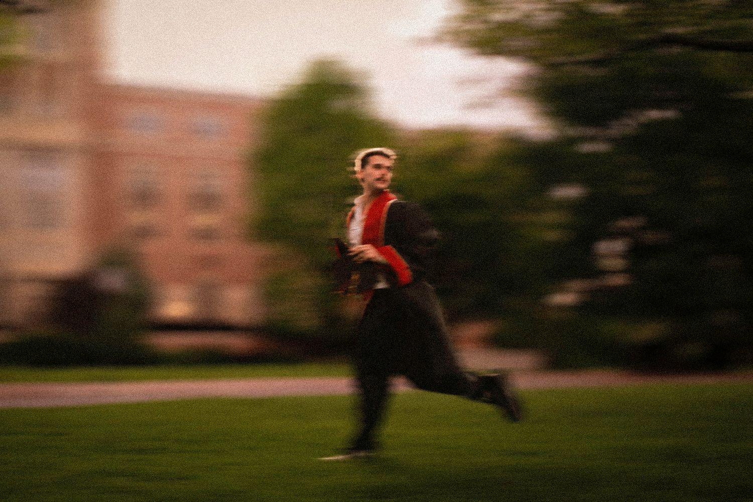 Graduate in cap and gown running across a green campus lawn with motion blur effect.