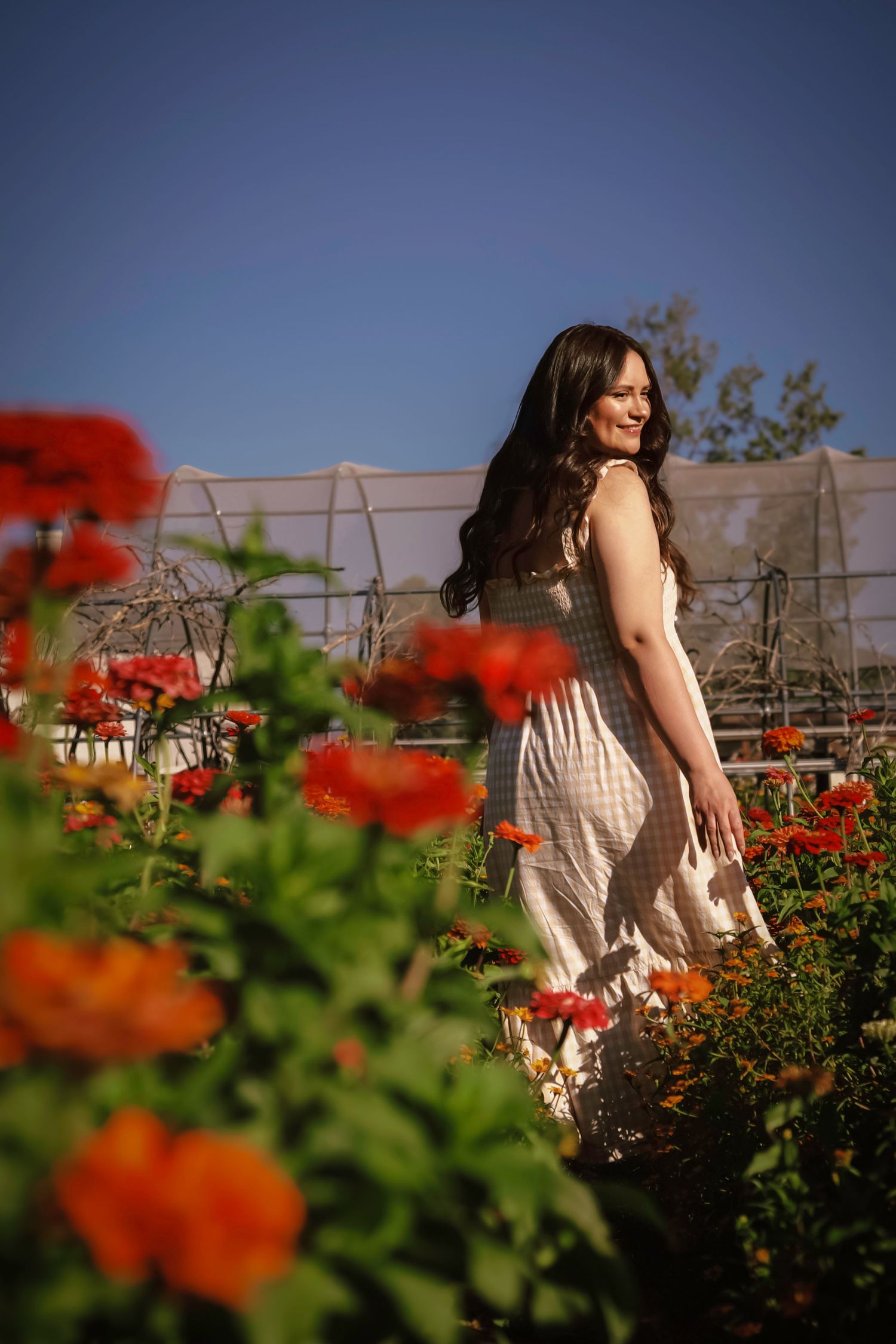 Smiling woman in white dress walking among vibrant red and orange flowers in a sunny garden.