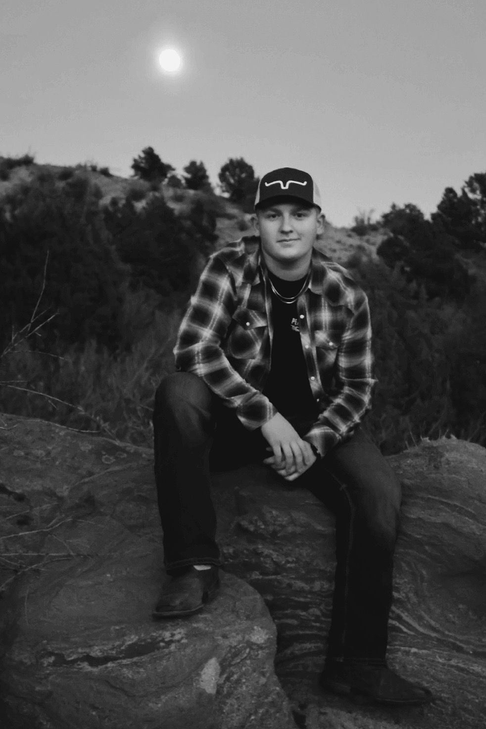 Black and white photo of young man in flannel sitting on a rock at dusk, full moon glowing in the sky behind him.