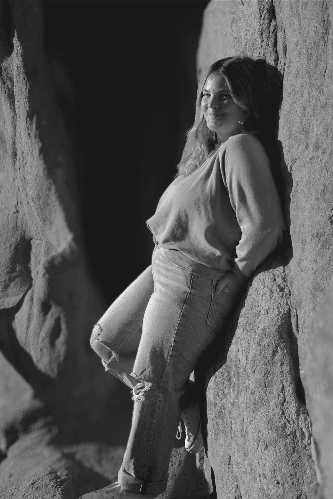 Young woman in ripped jeans leans against rock wall in dramatic lighting. Editorial-style black and white portrait.