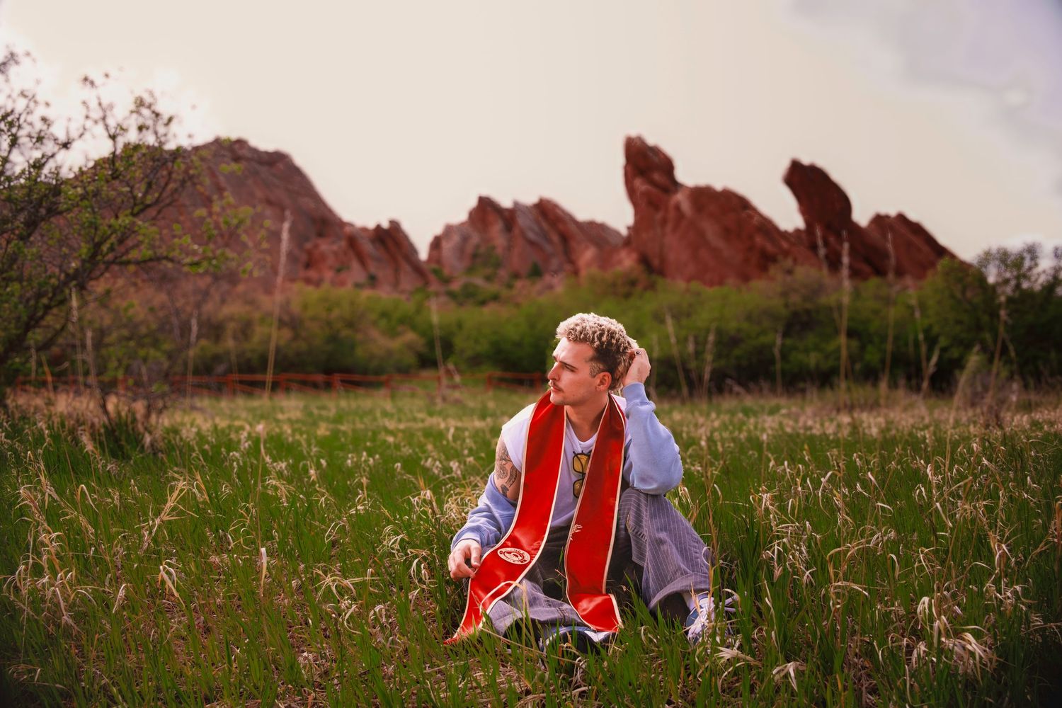 Graduate in cap and gown sitting in green field with red rock formations in background at sunset.