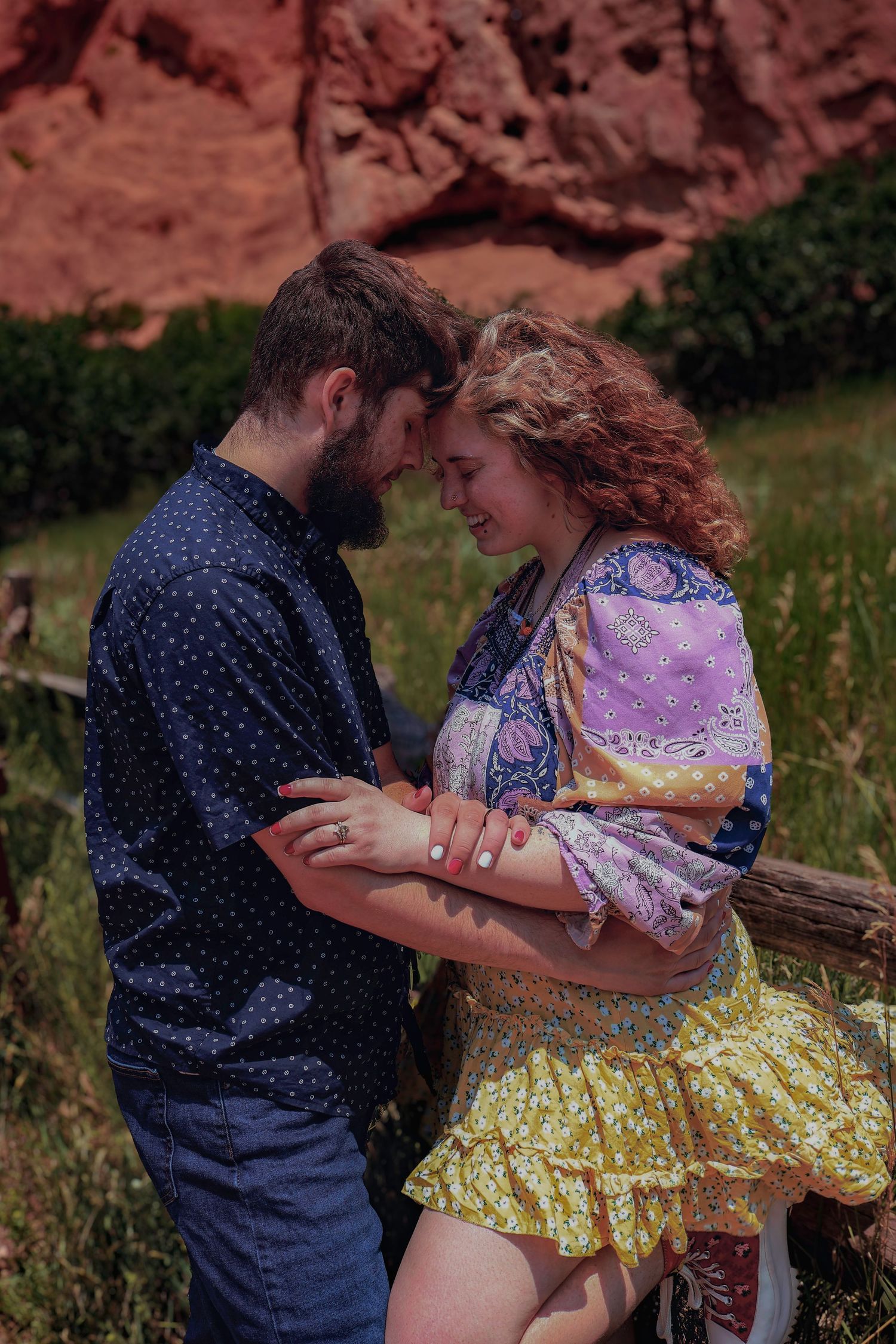 A couple shares a romantic moment outdoors near red rock formations, with the woman in a colorful bohemian dress.