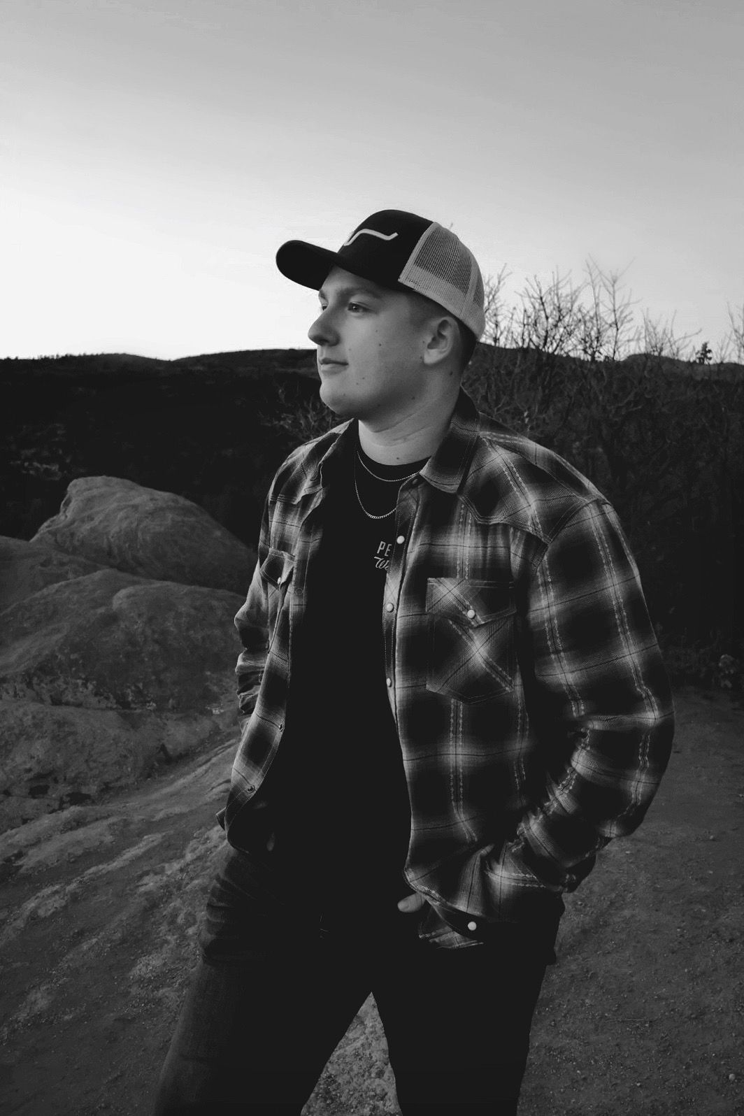 Young man in flannel and cap looks into distance on a mountain trail at sunset. Outdoor senior portrait photography in Colorado.