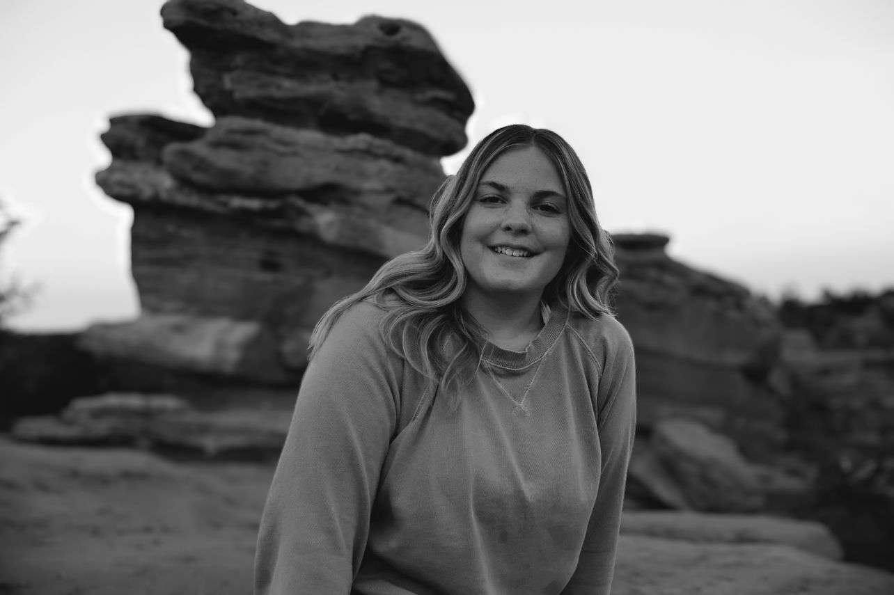 Black and white portrait of a woman smiling while seated on red rock formations during a moody golden hour photo shoot.