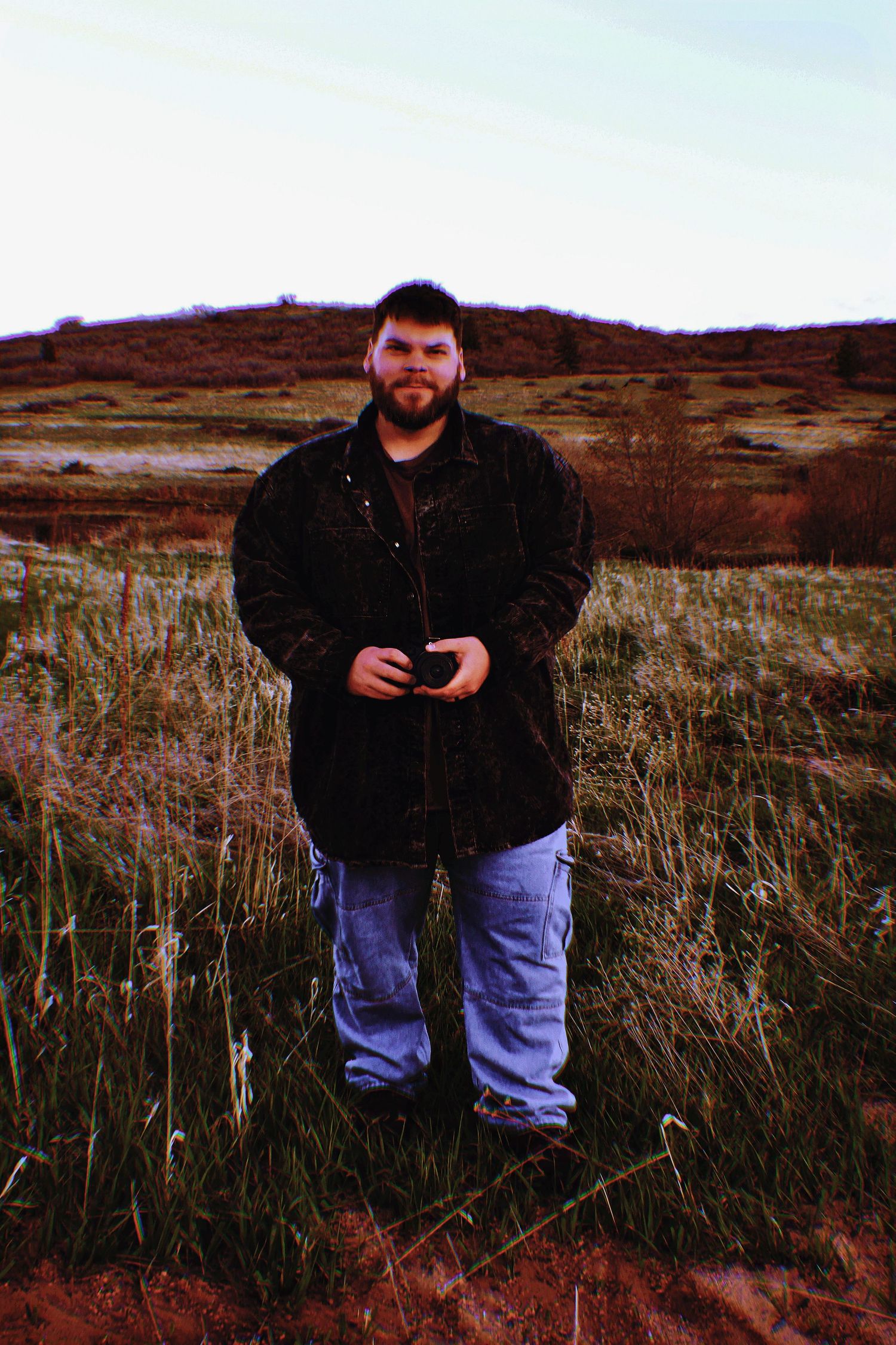 A bearded man in a dark jacket and jeans stands in a rural field with hills in the background at Forest Lakes, in Monument, CO.