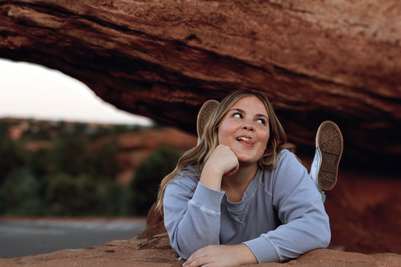 Teen girl laying playfully on red rocks with legs raised. Candid lifestyle senior photo in Colorado outdoors.