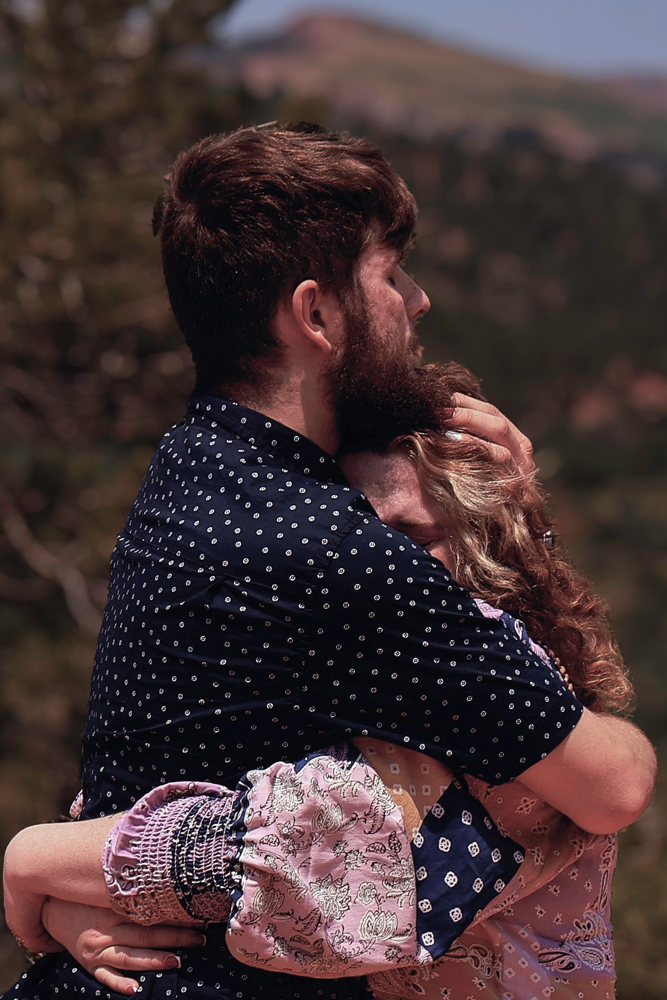 Bearded man in polka dot shirt embracing a woman with curly hair outdoors with mountain scenery.