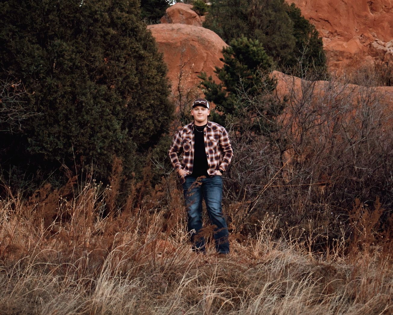 Young man standing in tall grass with red rocks in background, wearing flannel during a rustic senior portrait session.