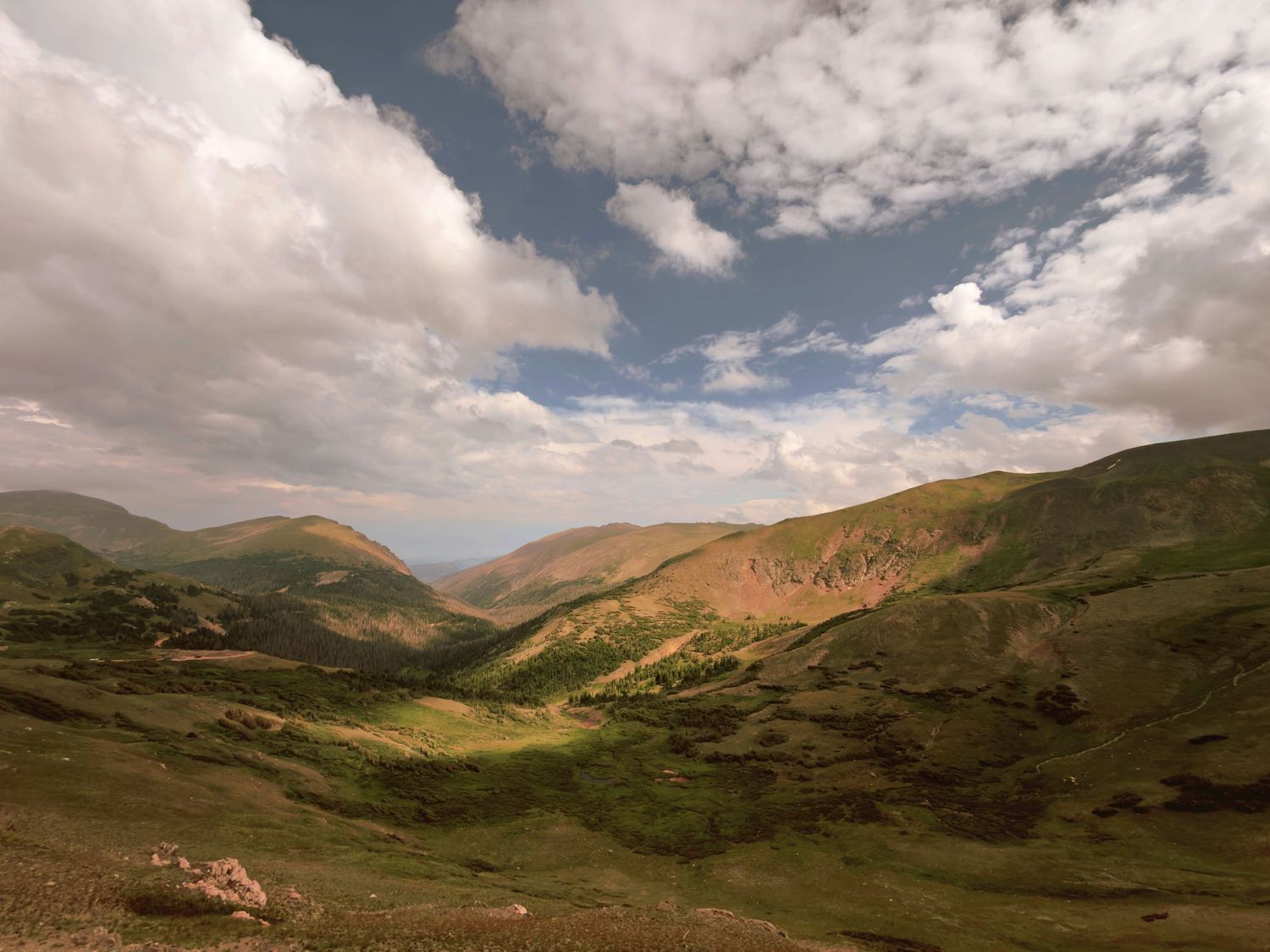 Lush green mountain valley with rolling hills under a dramatic cloudy blue sky in a highland landscape in Rocky Mountain National Park.