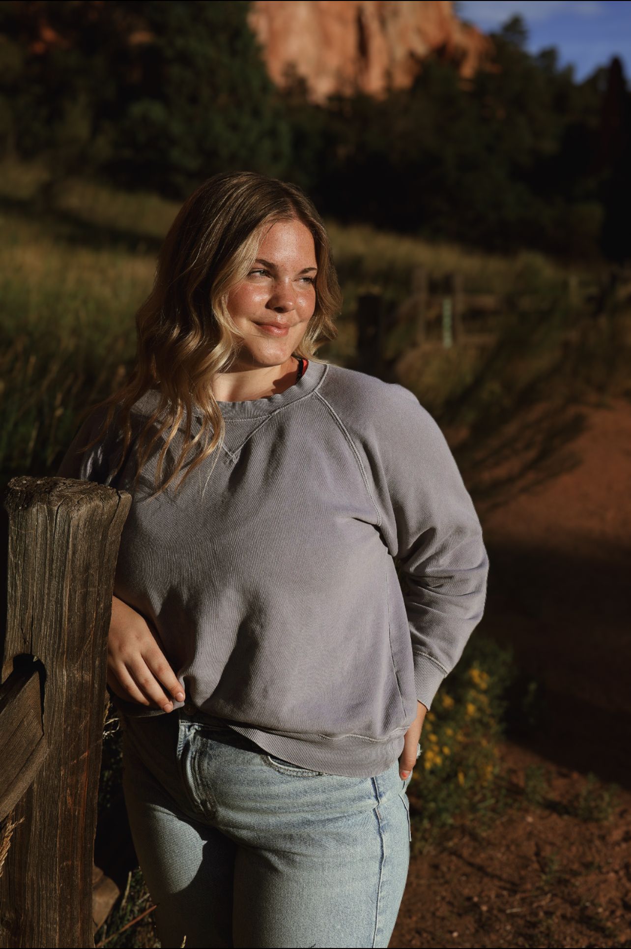 Girl in purple top leans on wooden fence, smiling in golden hour light. Garden of the Gods senior session.