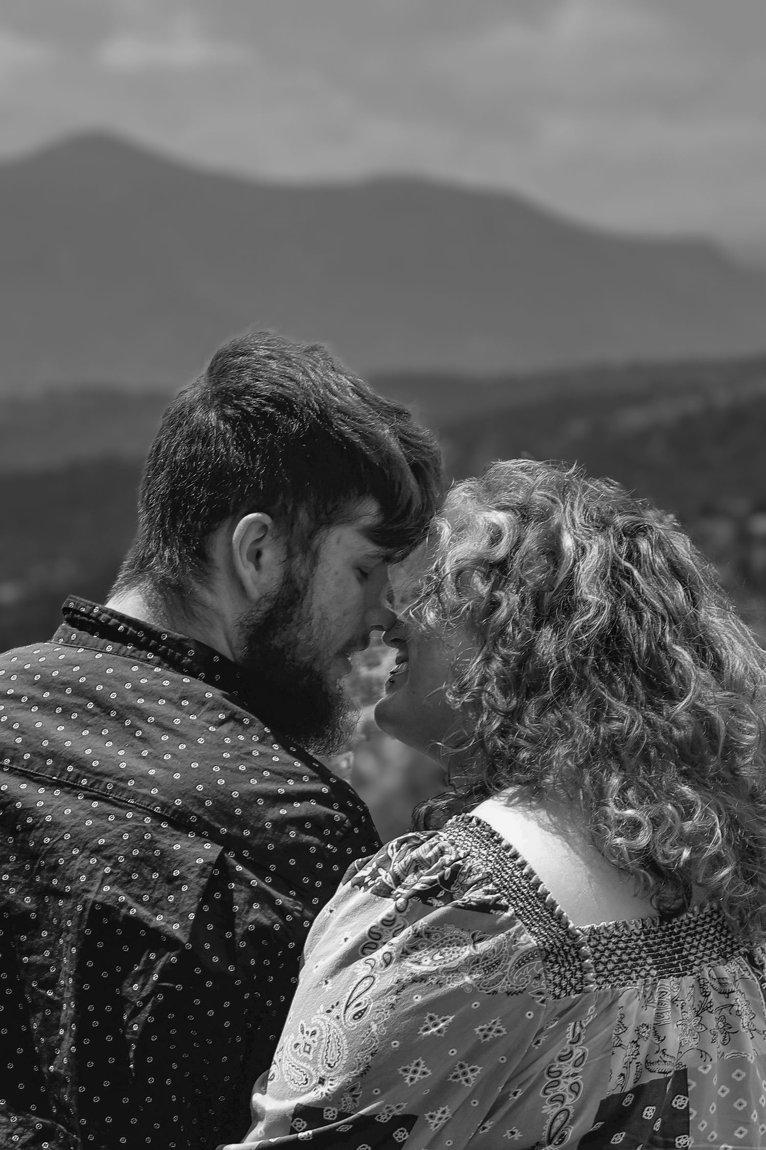 Black and white photo of a romantic couple leaning foreheads together with mountain landscape in background.